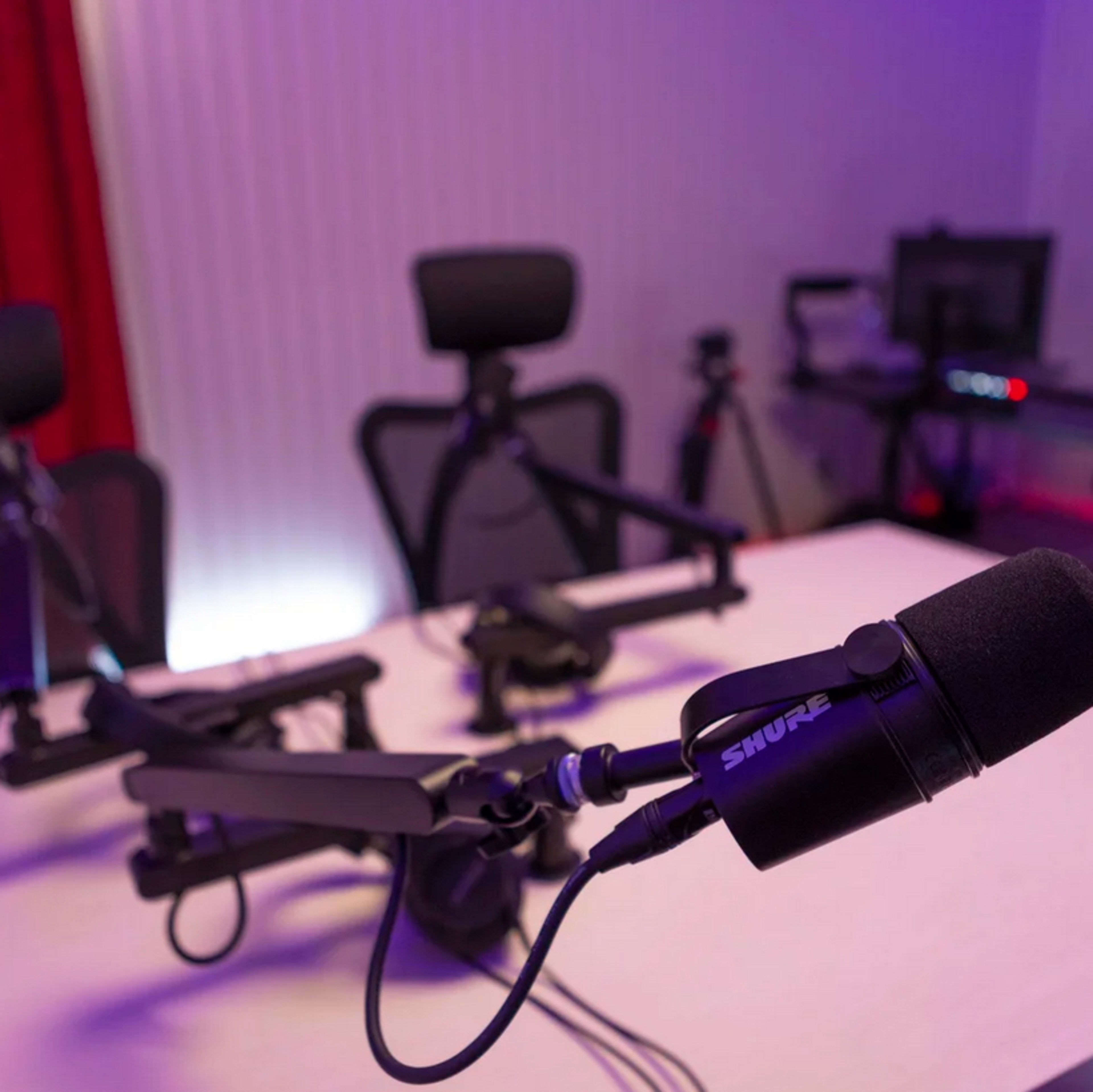 The image shows a podcasting setup featuring a Shure microphone mounted on a boom arm, with several ergonomic chairs and equipment in the background.