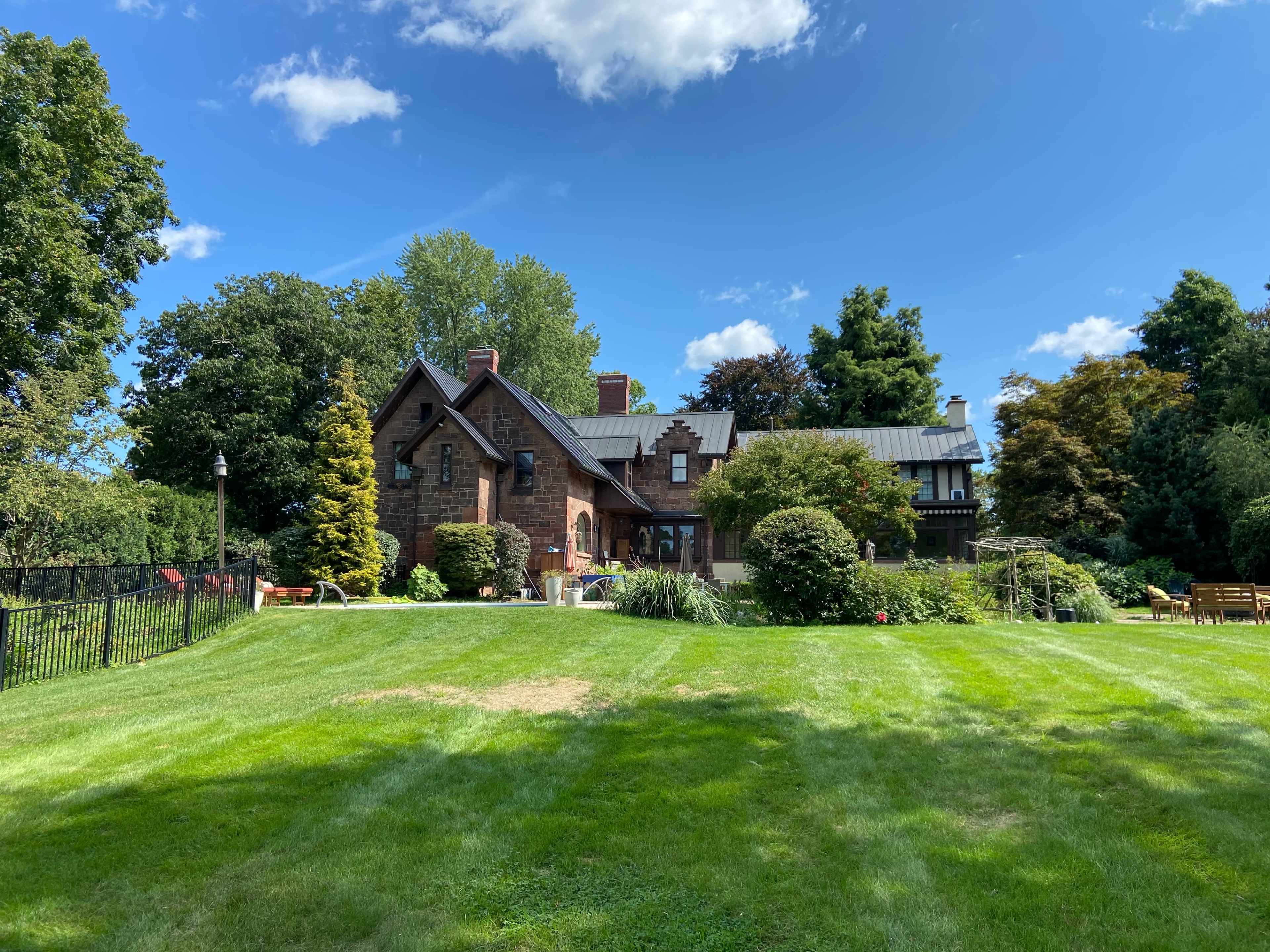 A large, two-story brick house is surrounded by a green lawn and various trees under a blue sky with a few clouds.