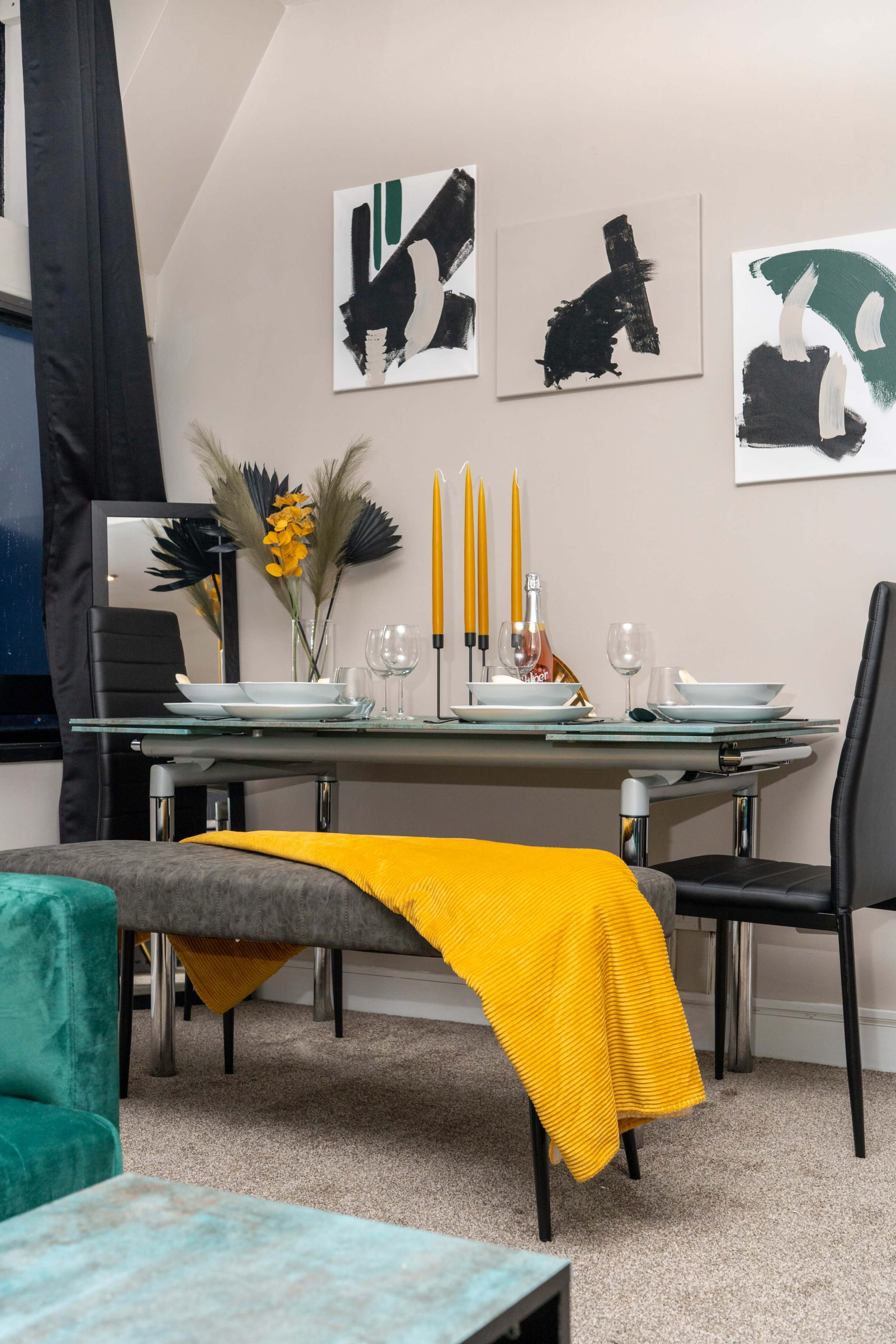 A modern dining setup features a glass table with plates, glasses, and tall yellow candles, accompanied by a bench draped with a yellow throw and artwork on the walls.