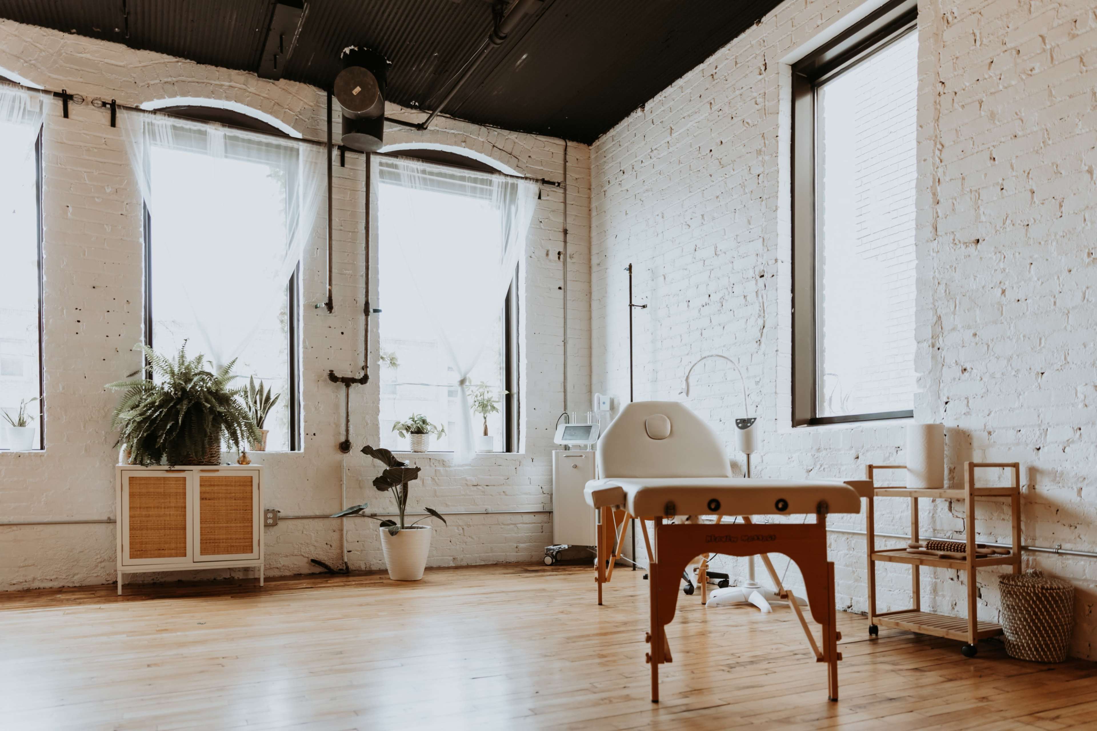 A bright, minimalistic treatment room featuring a massage table, large windows with sheer curtains, and plants arranged artistically.