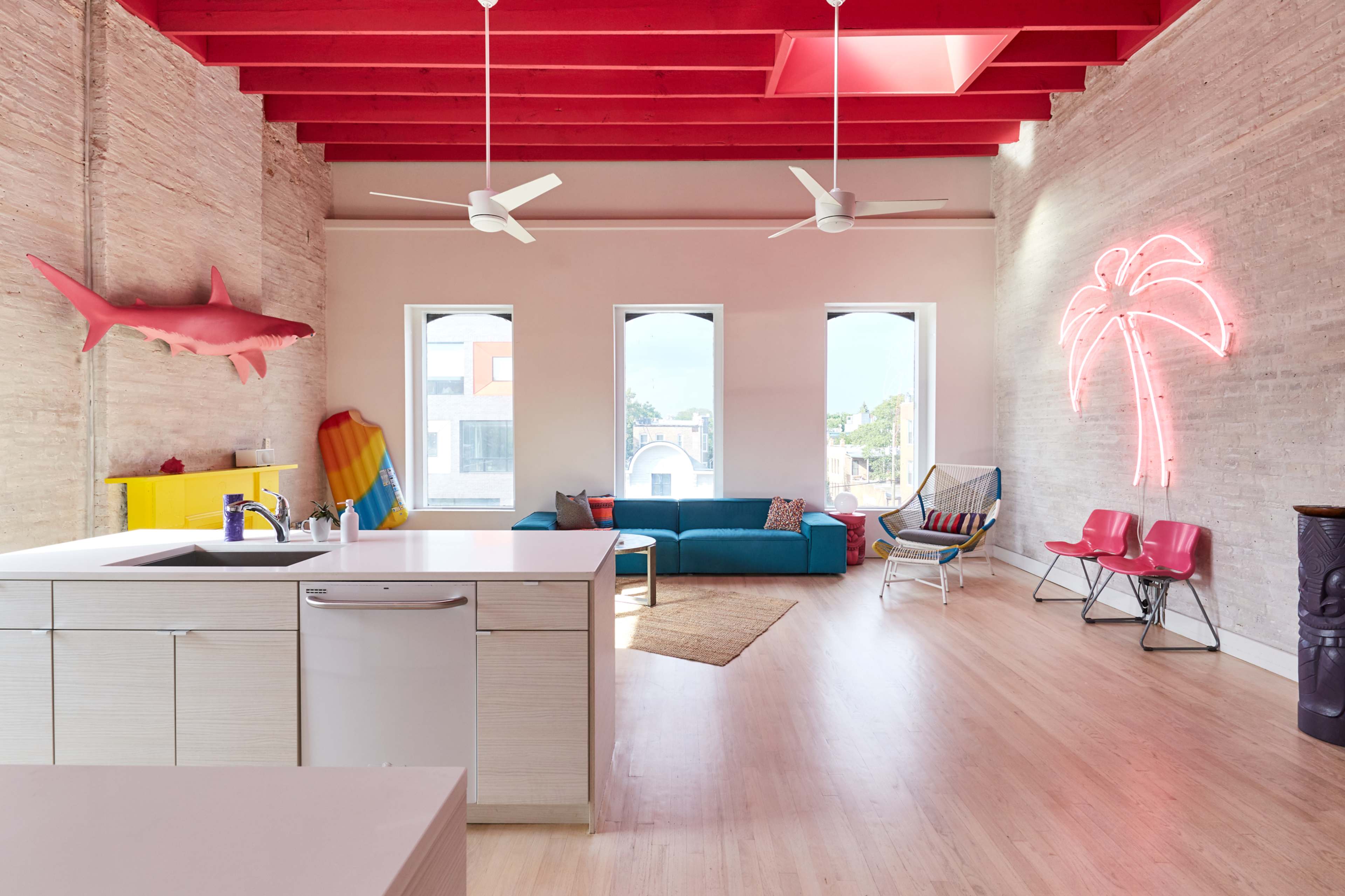A spacious, brightly lit living area features a modern kitchen with a white island, red ceiling beams, and colorful decor including a shark sculpture and neon palm tree.