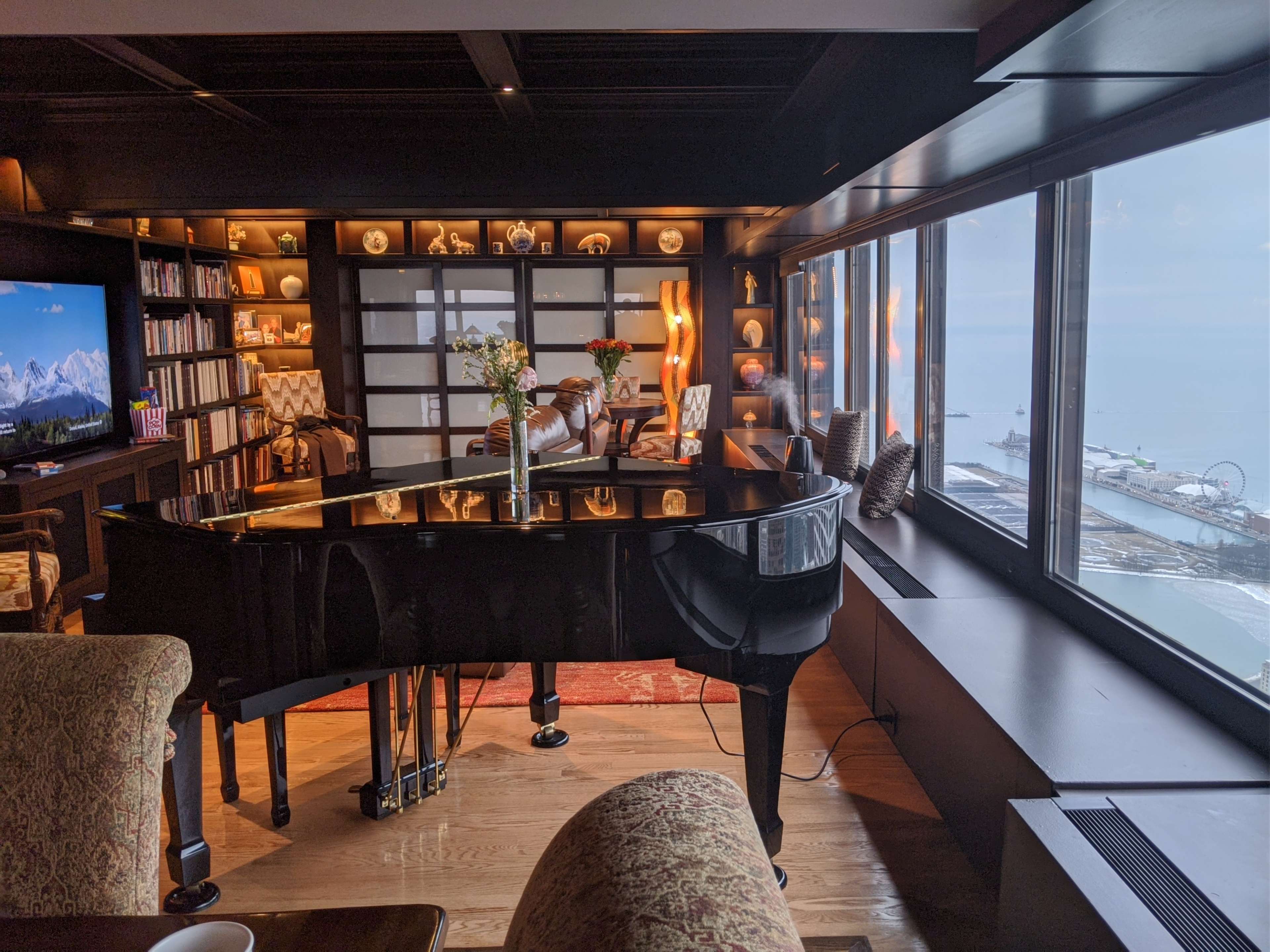 A grand piano sits in a stylishly decorated living room with large windows overlooking a seaside view.