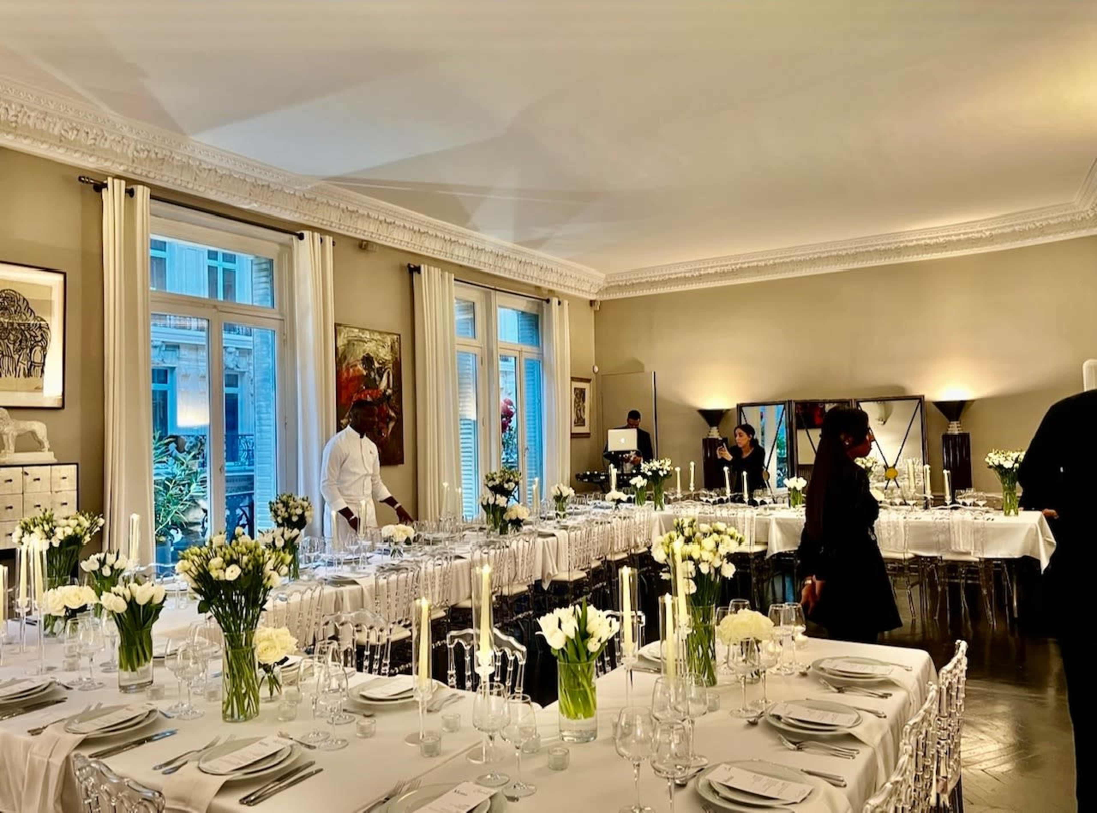 A elegantly arranged dining room features a long table set with white flowers, candles, and glassware, as staff prepare for an event.