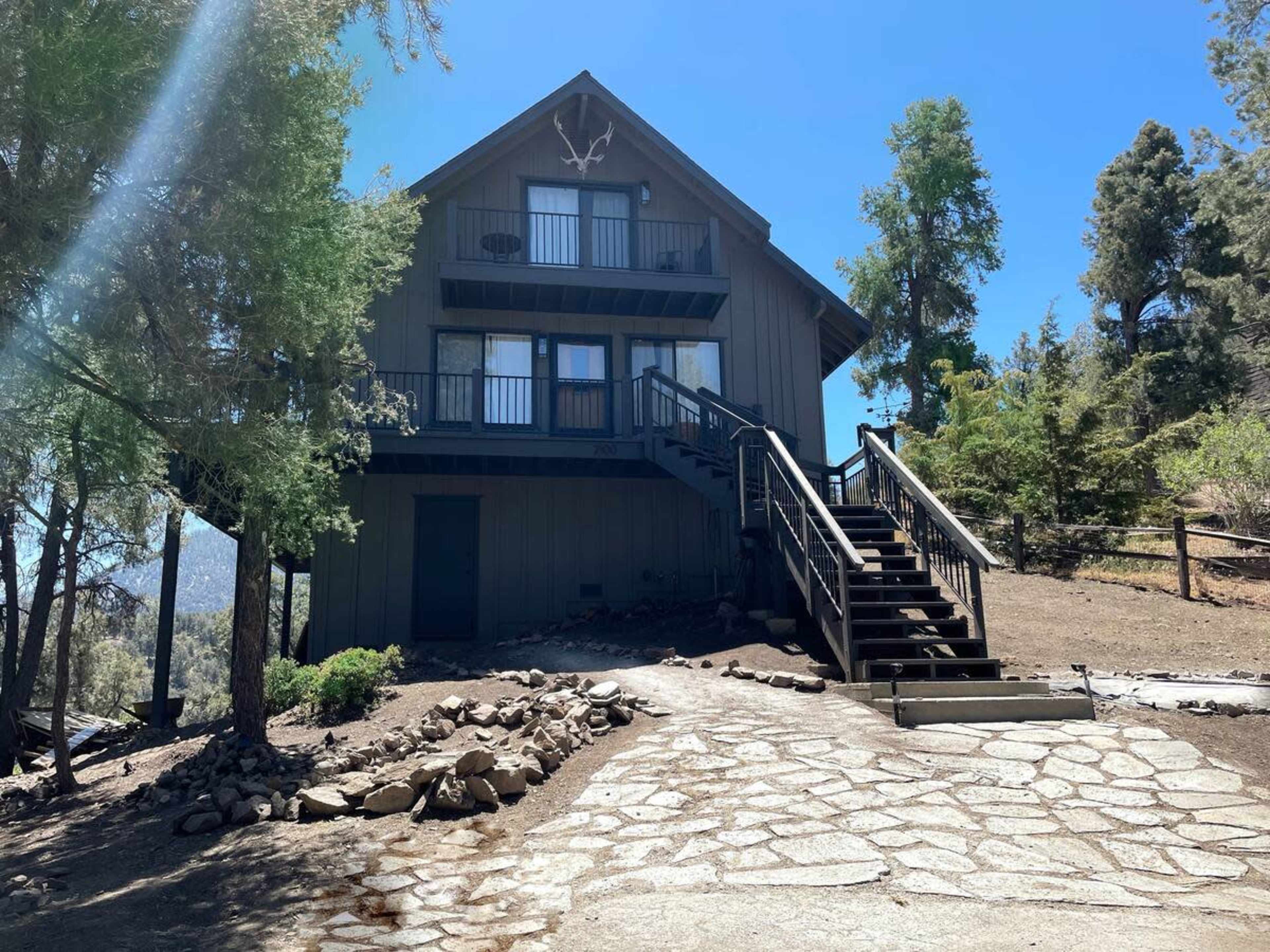 A three-story cabin with a wooden exterior and a set of stairs leads up to a covered porch surrounded by trees.