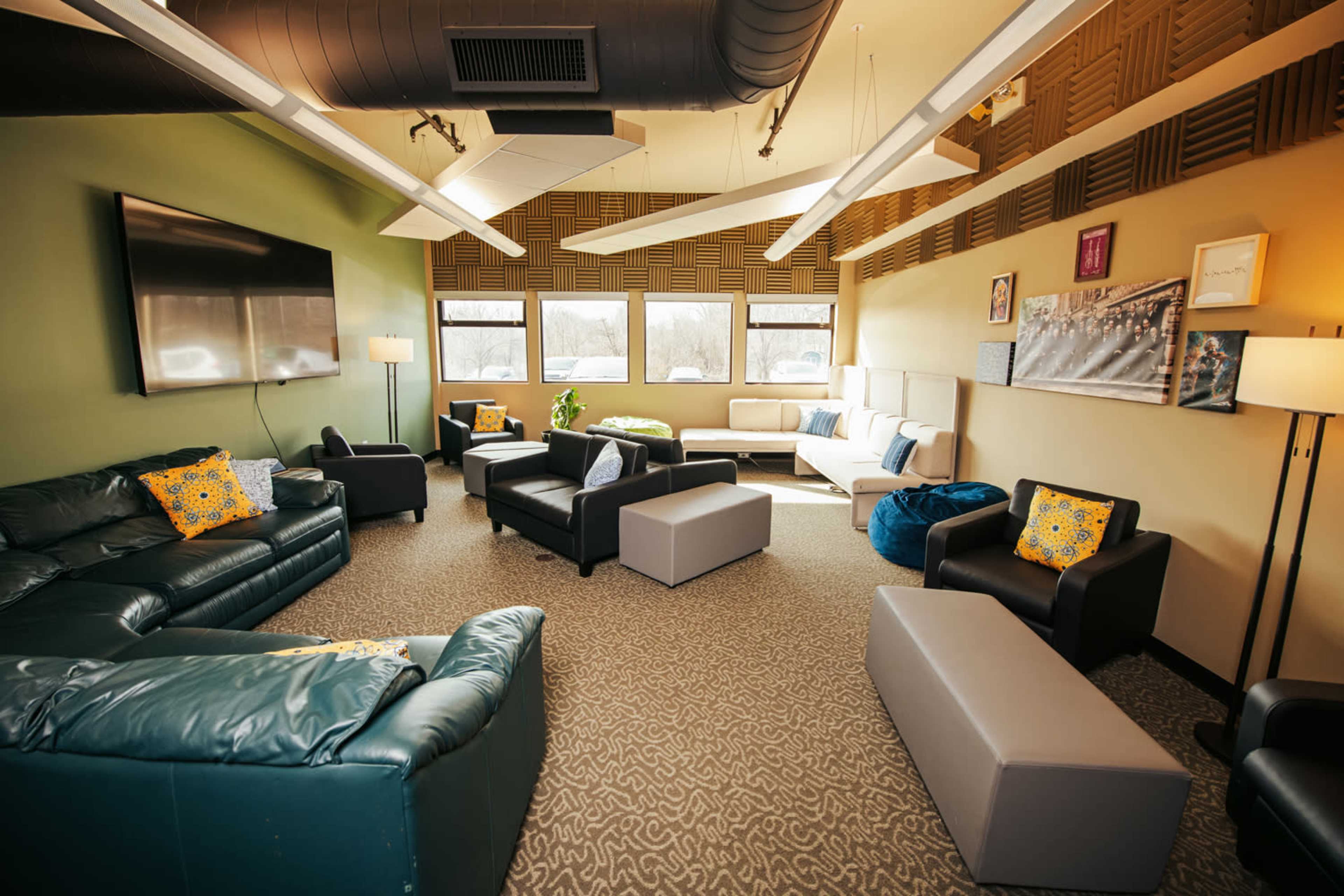 The image shows a spacious lounge area with several couches, armchairs, and tables arranged around a central space, featuring windows that let in natural light.