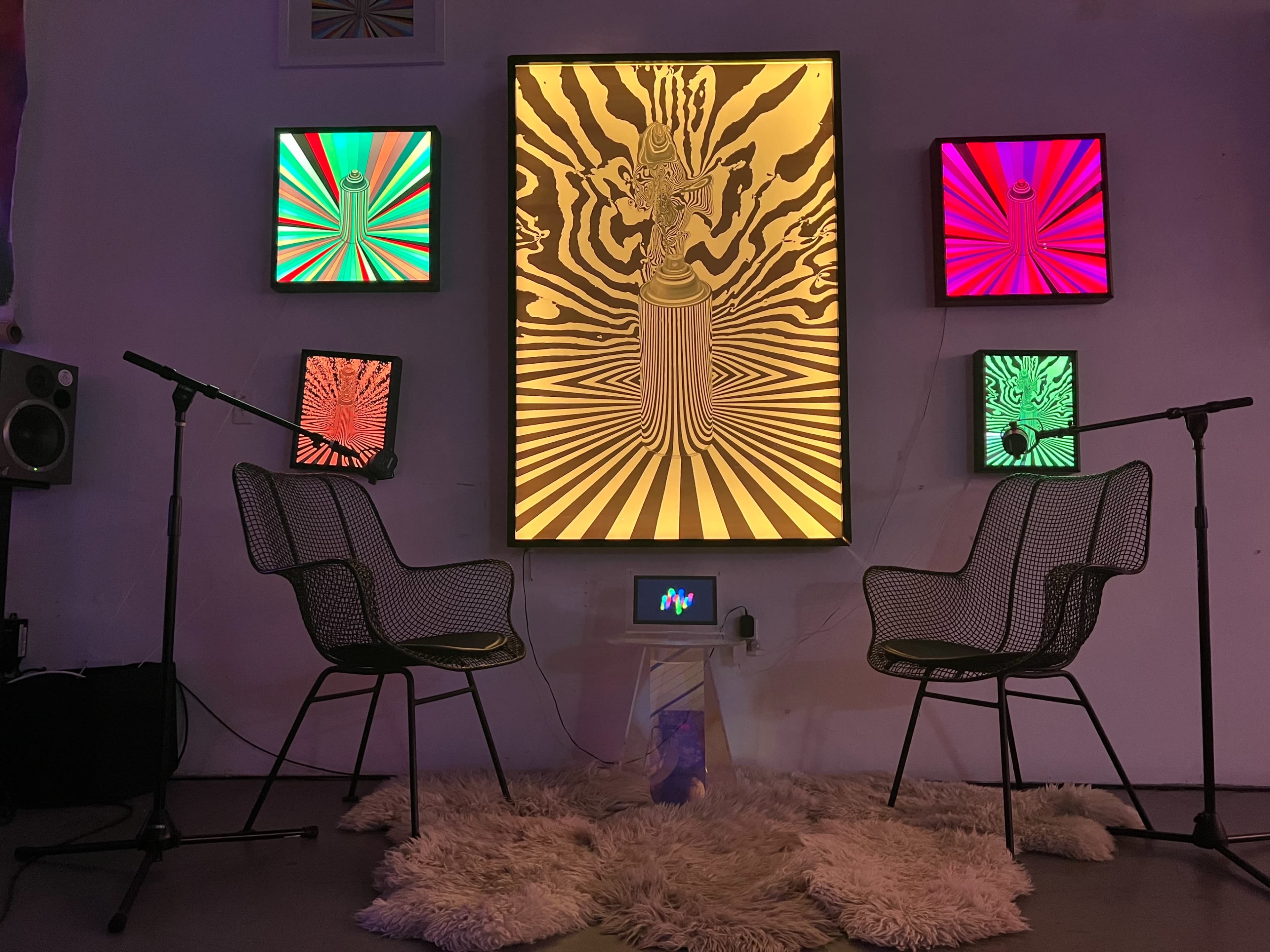 A colorful art installation featuring a central illuminated piece with a design of a figure, flanked by smaller vibrant screens, with two wire chairs facing a small display on a rug.