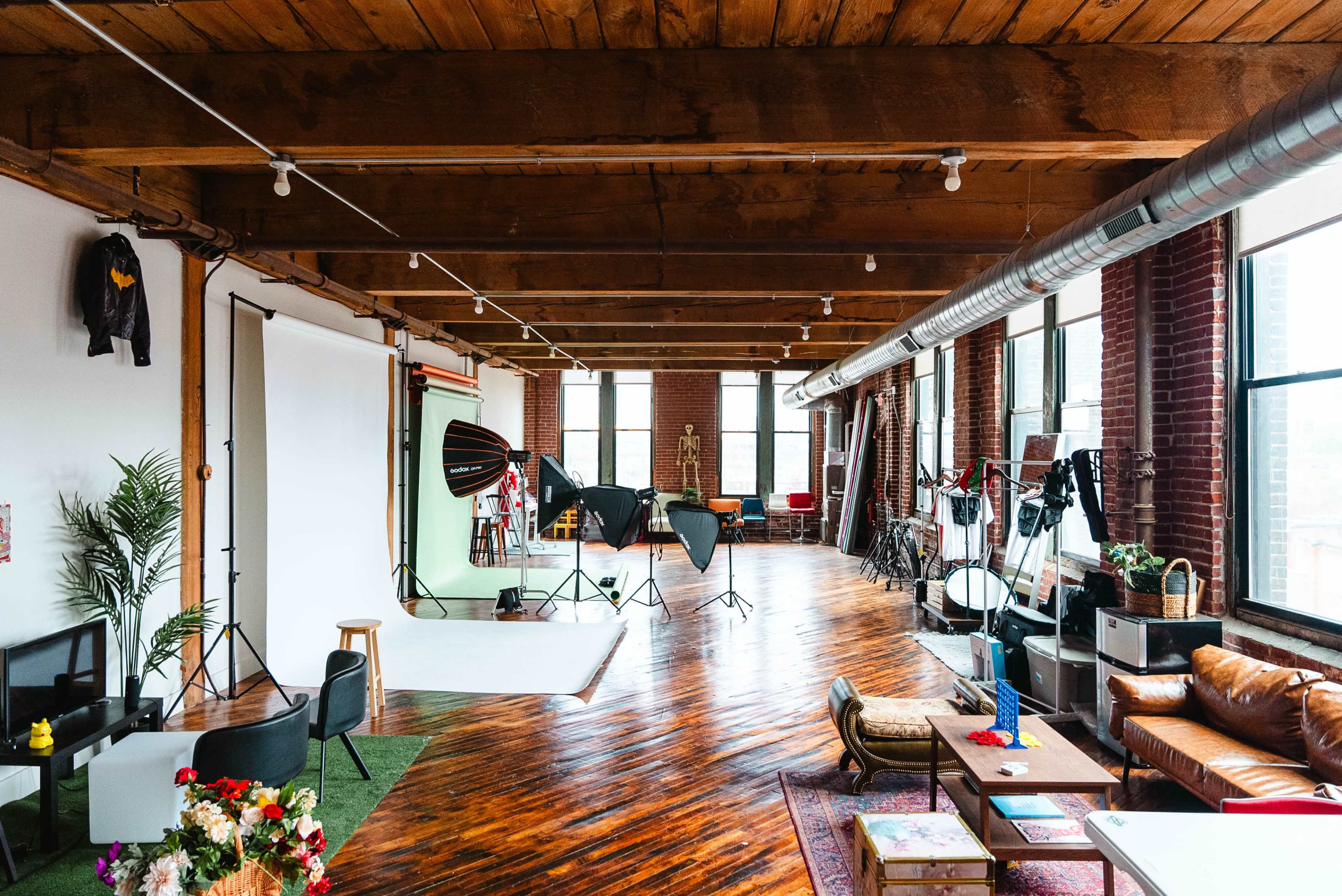 A spacious photo studio with wooden floors, large windows, and various lighting equipment set up around a backdrop.