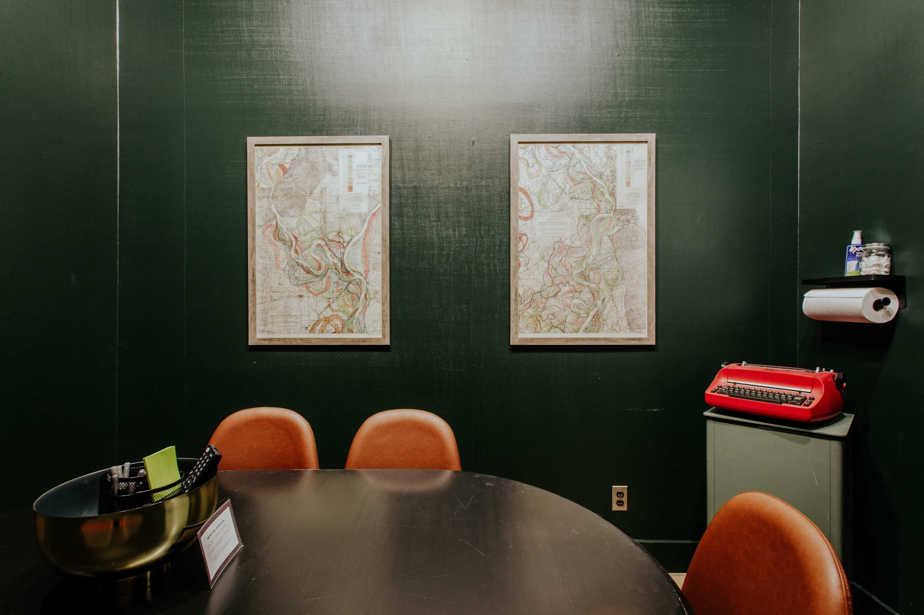 A small room with dark green walls, featuring two framed maps on the walls, a round black table, four brown chairs, and a red typewriter on a green filing cabinet.
