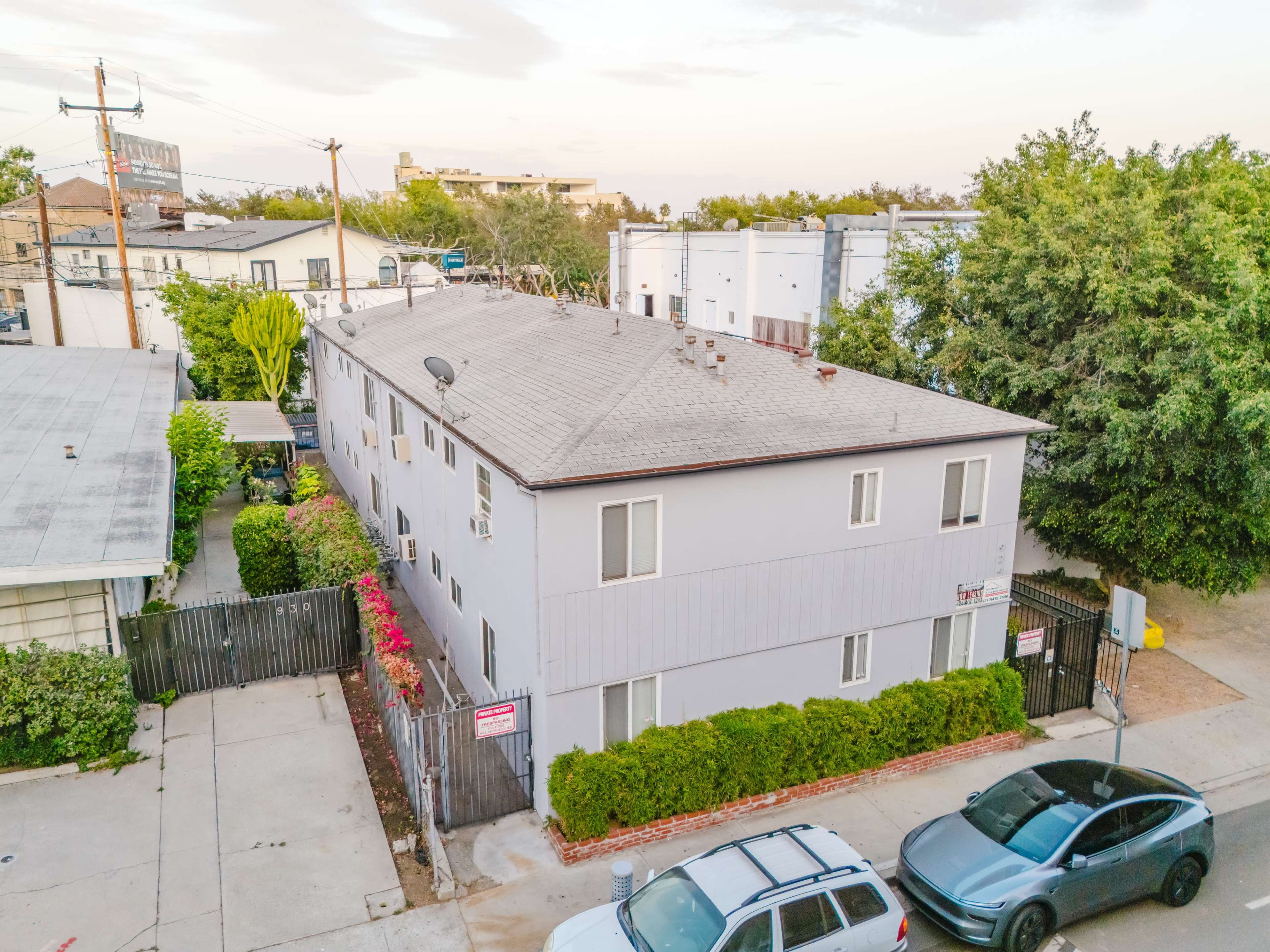The image shows a two-story residential building surrounded by greenery and parked cars.