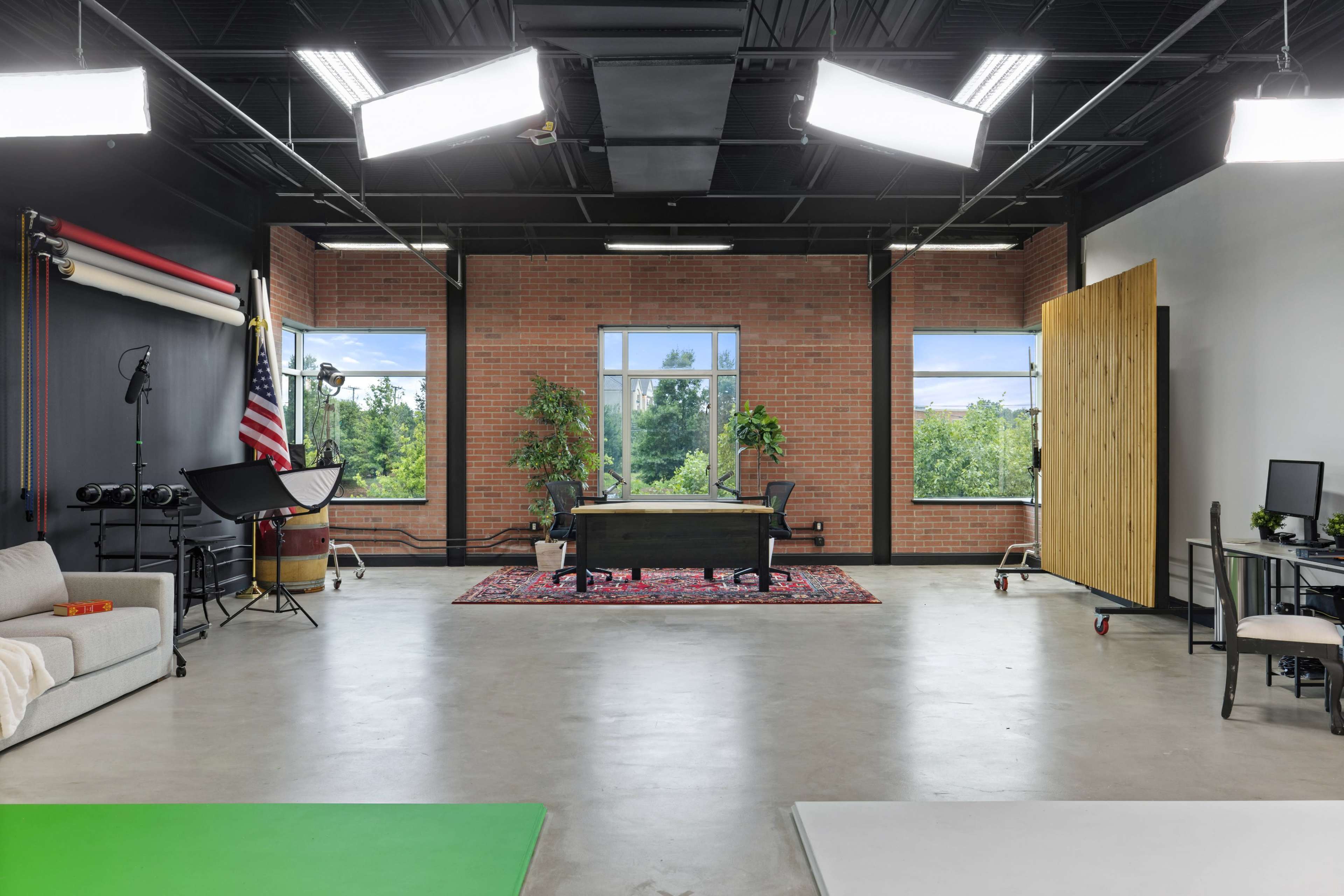 A spacious studio with large windows, a wooden desk at the center, seating areas, and various lighting equipment.