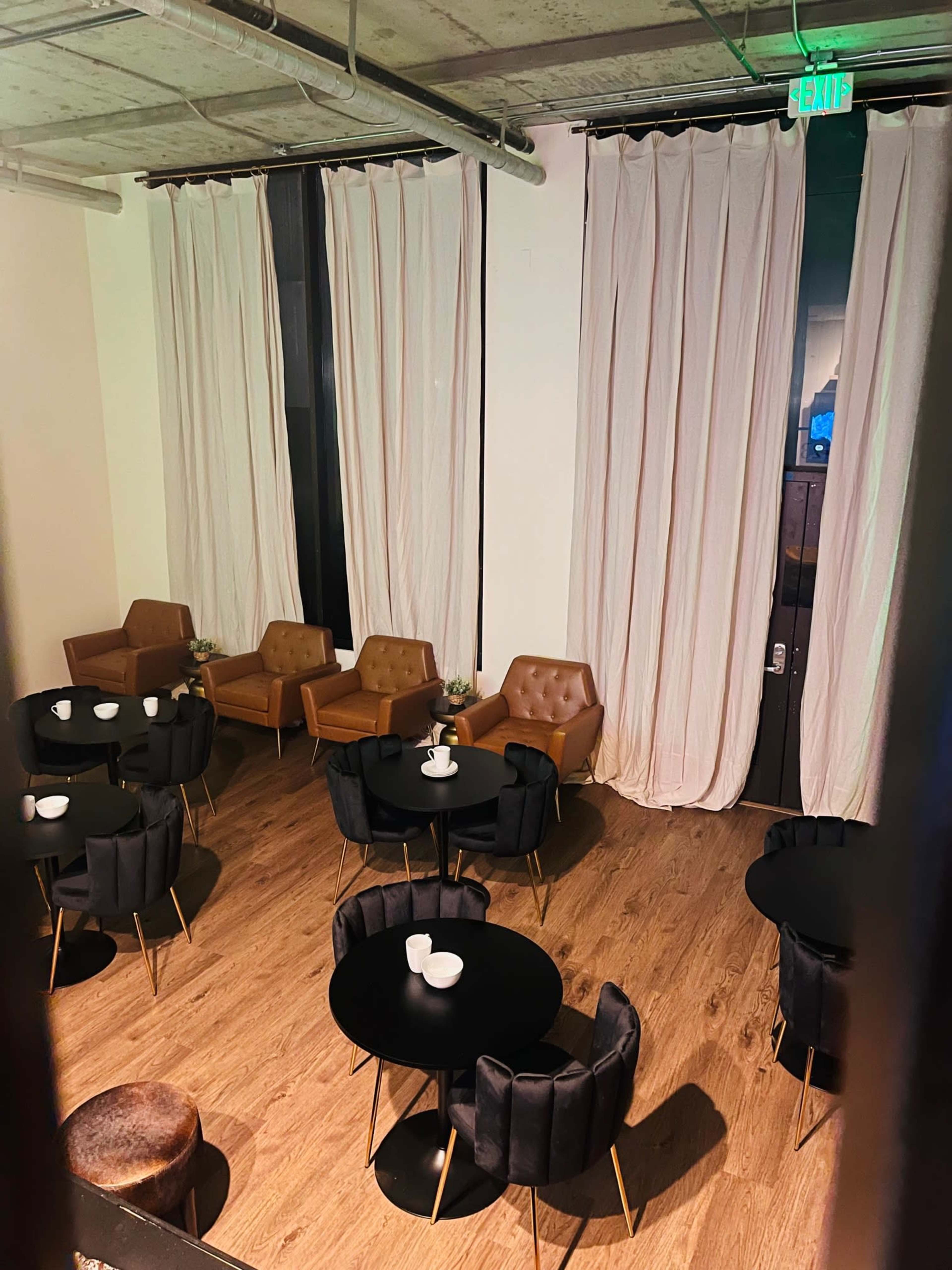 A top-down view of a modern lounge area with several black tables surrounded by brown and black upholstered chairs, and floor-to-ceiling curtains on the windows.