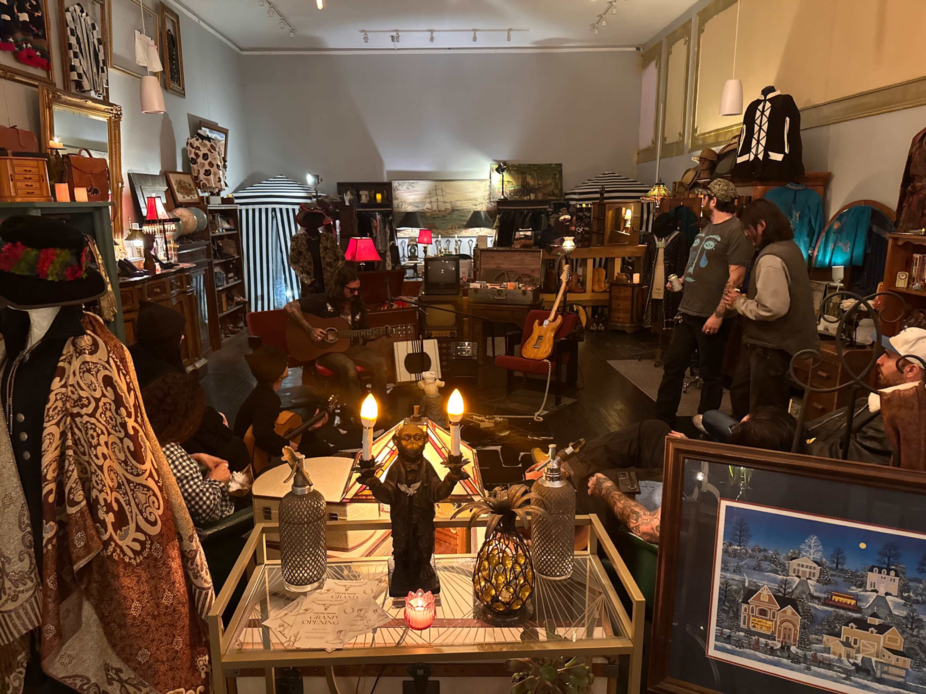 The image shows a vintage shop filled with various clothing items, guitars, and decorative objects, with several people engaged in conversation and activities around a central seating area.