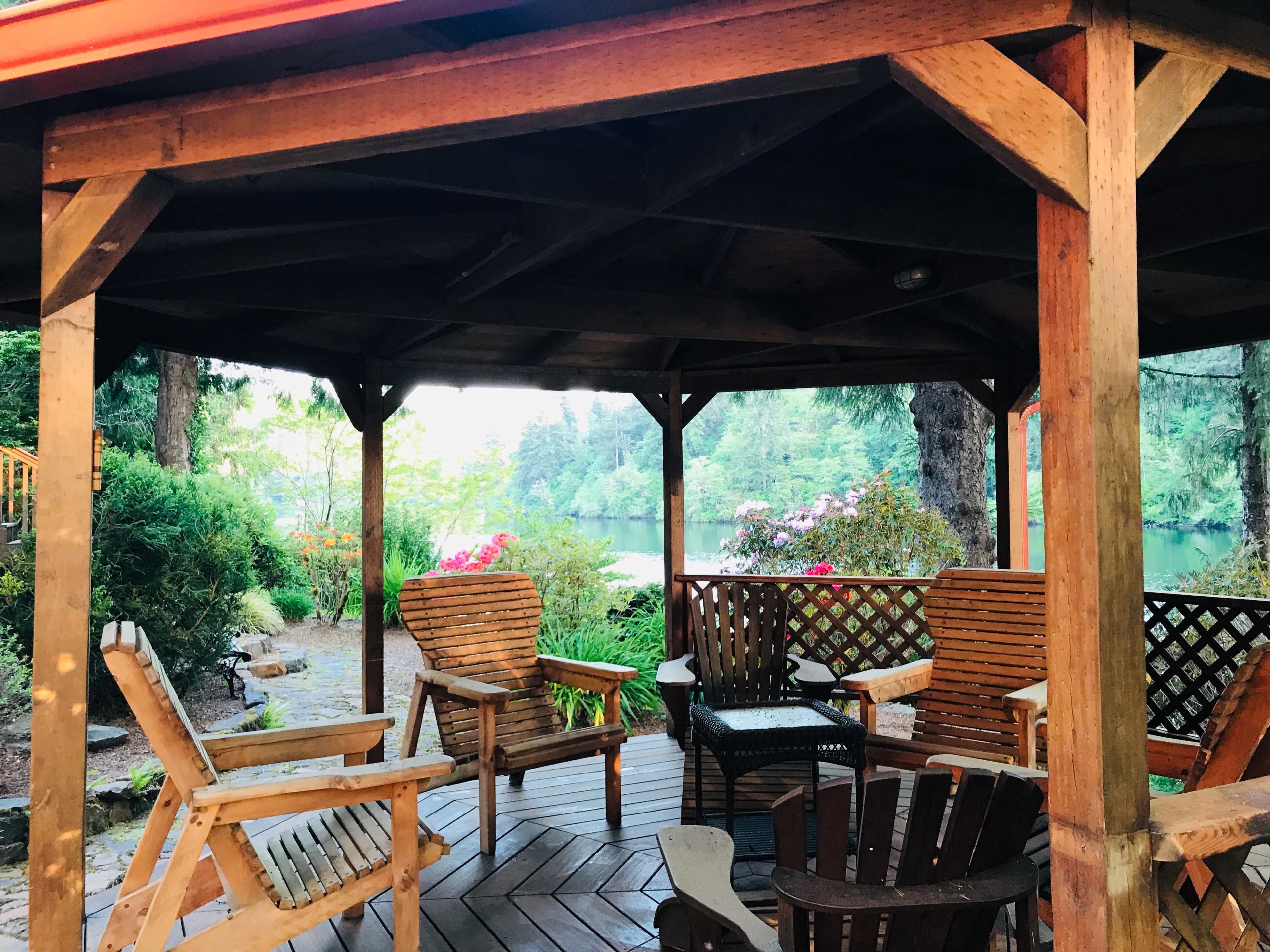 A wooden gazebo with several chairs overlooks a calm lake surrounded by trees and blooming flowers.