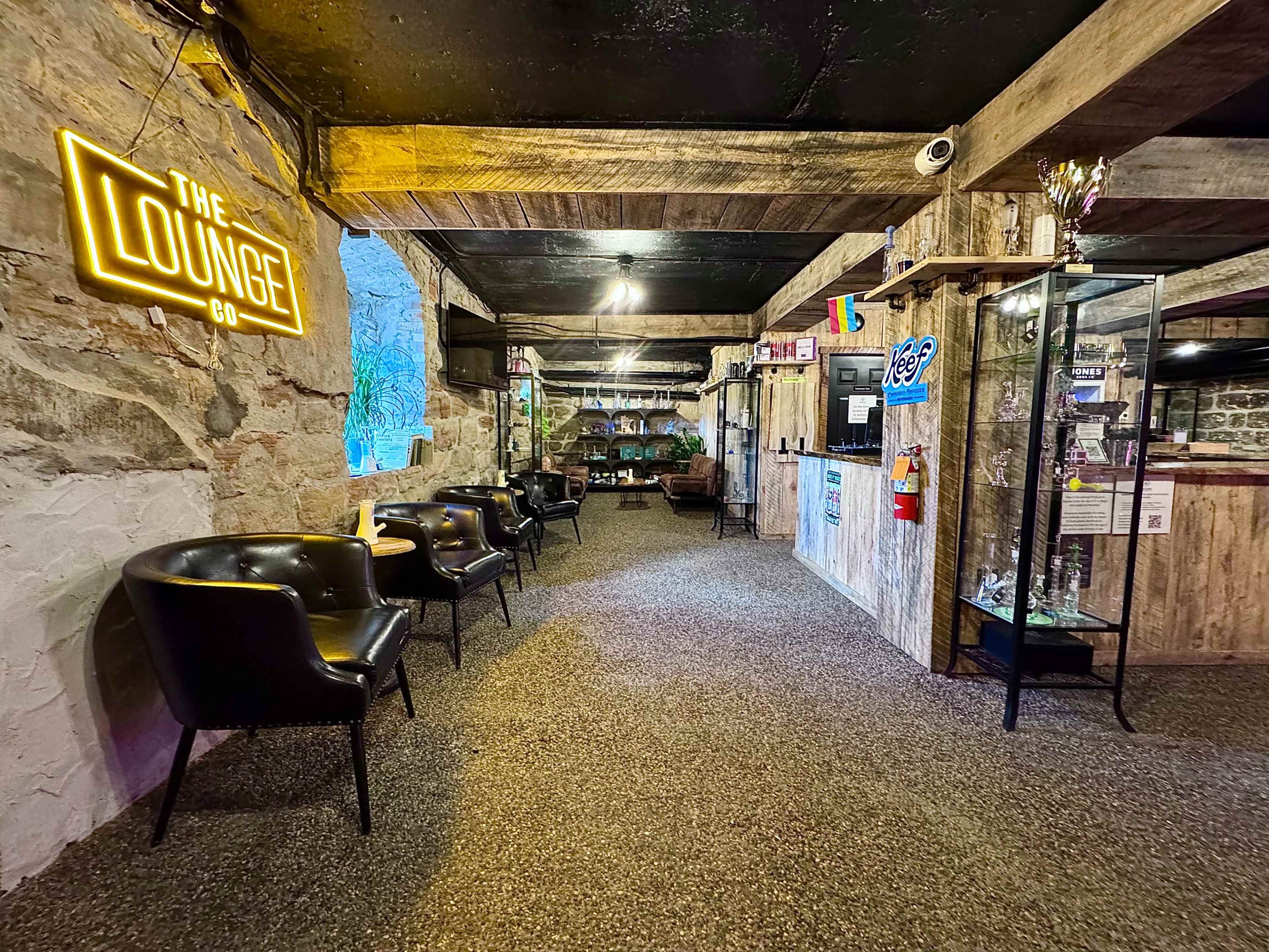 The image shows a cozy lounge area with stone walls, wooden beams, and a neon sign, featuring black chairs along a gravel floor and various decorative items in glass displays.