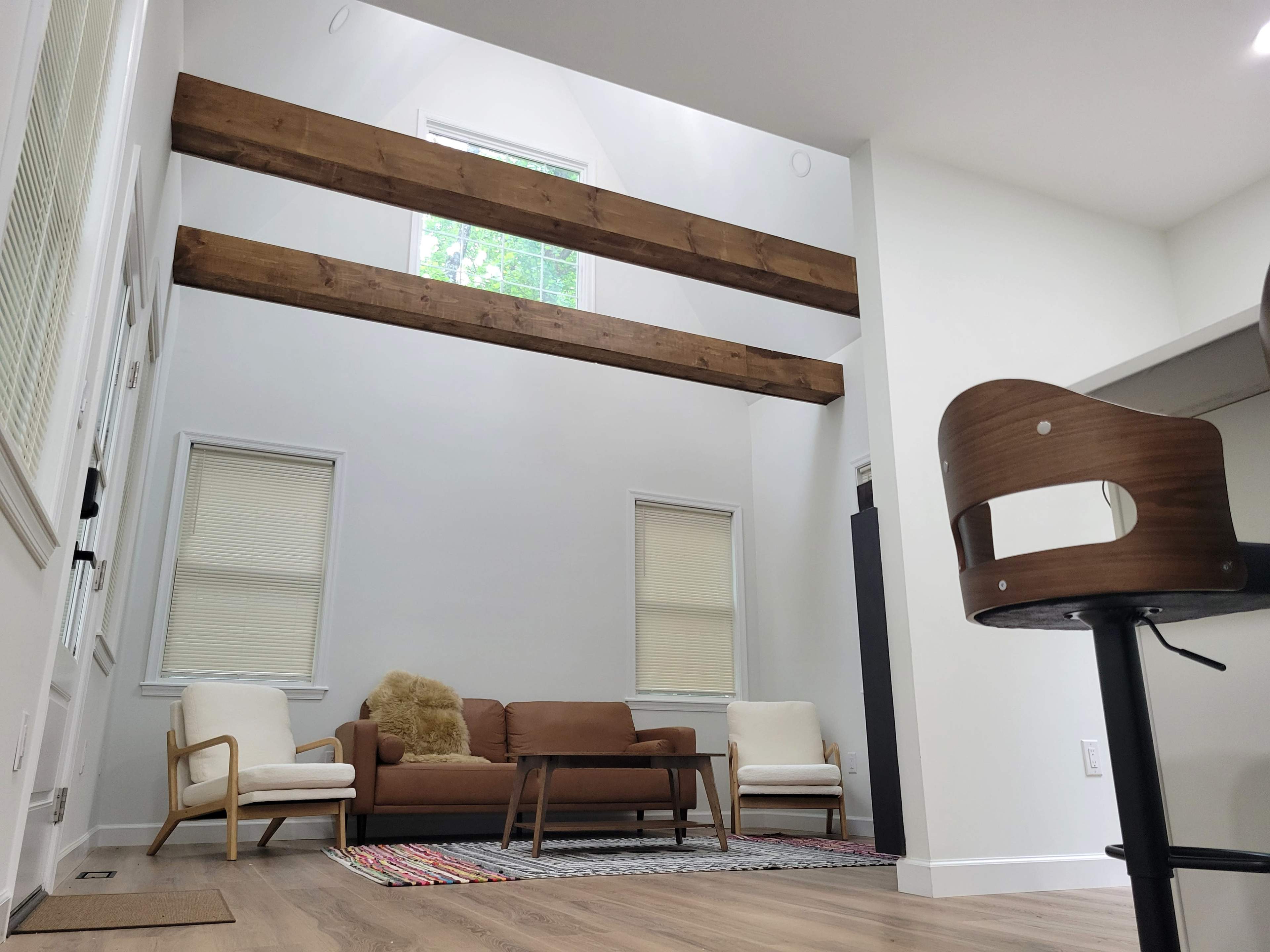 The image shows a modern living room with high ceilings, featuring wooden beams, a brown sofa, two armchairs, and a rug on a hardwood floor.
