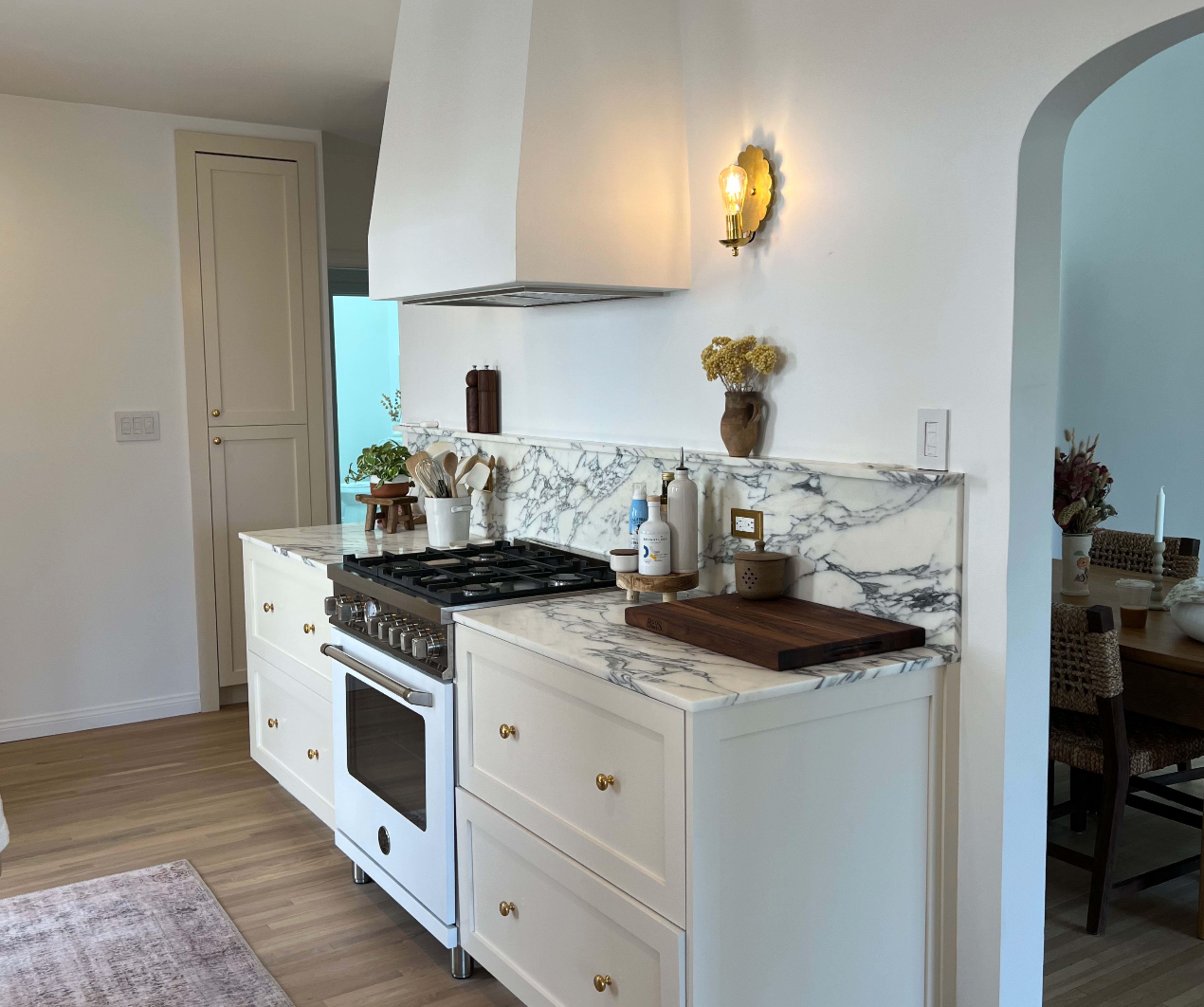 The image shows a modern kitchen with a marble countertop, white cabinets, and a gas stove beneath a chimney hood.