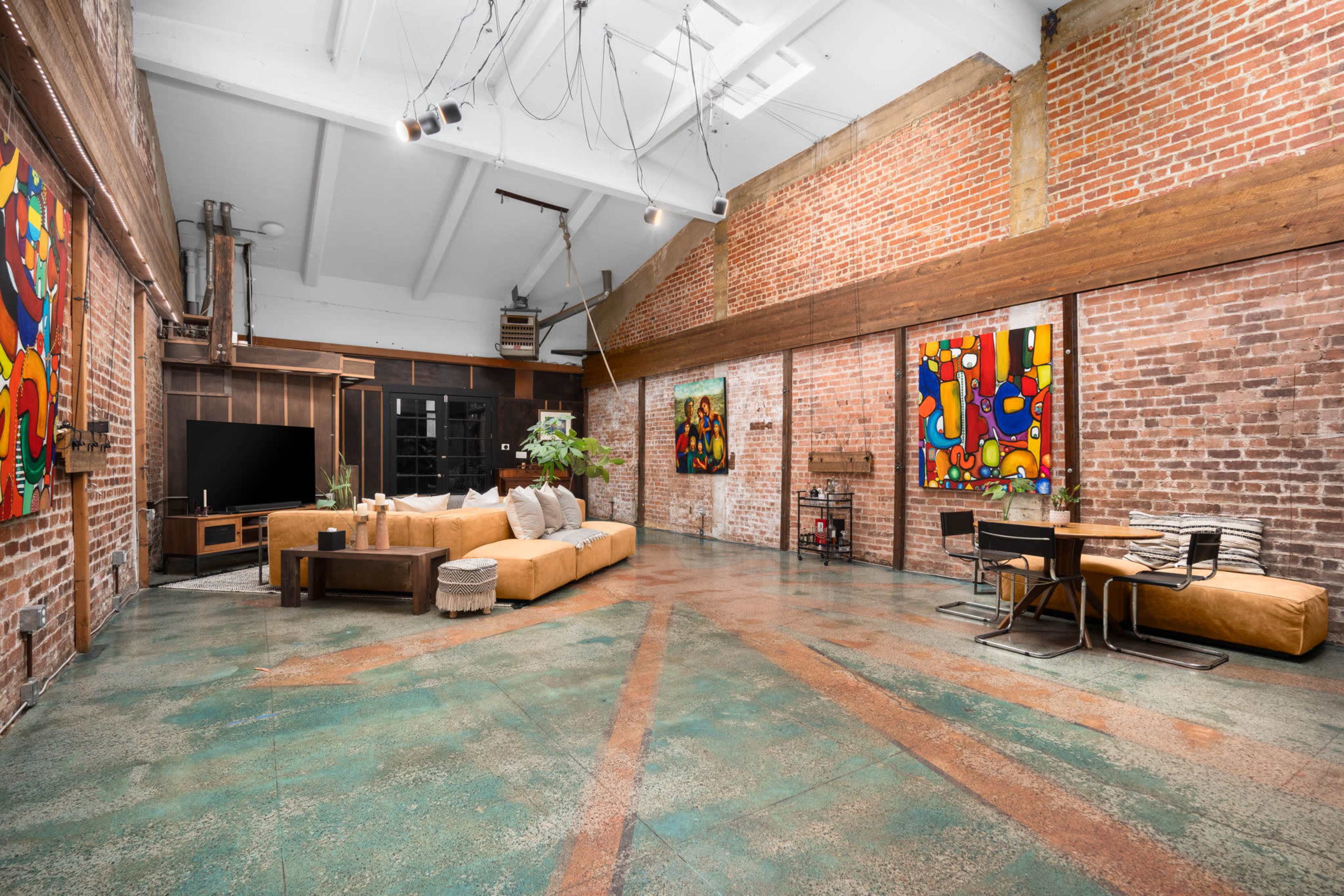 The spacious interior features exposed brick walls, a large sectional sofa, and colorful artwork, with a dining area and modern television setup.
