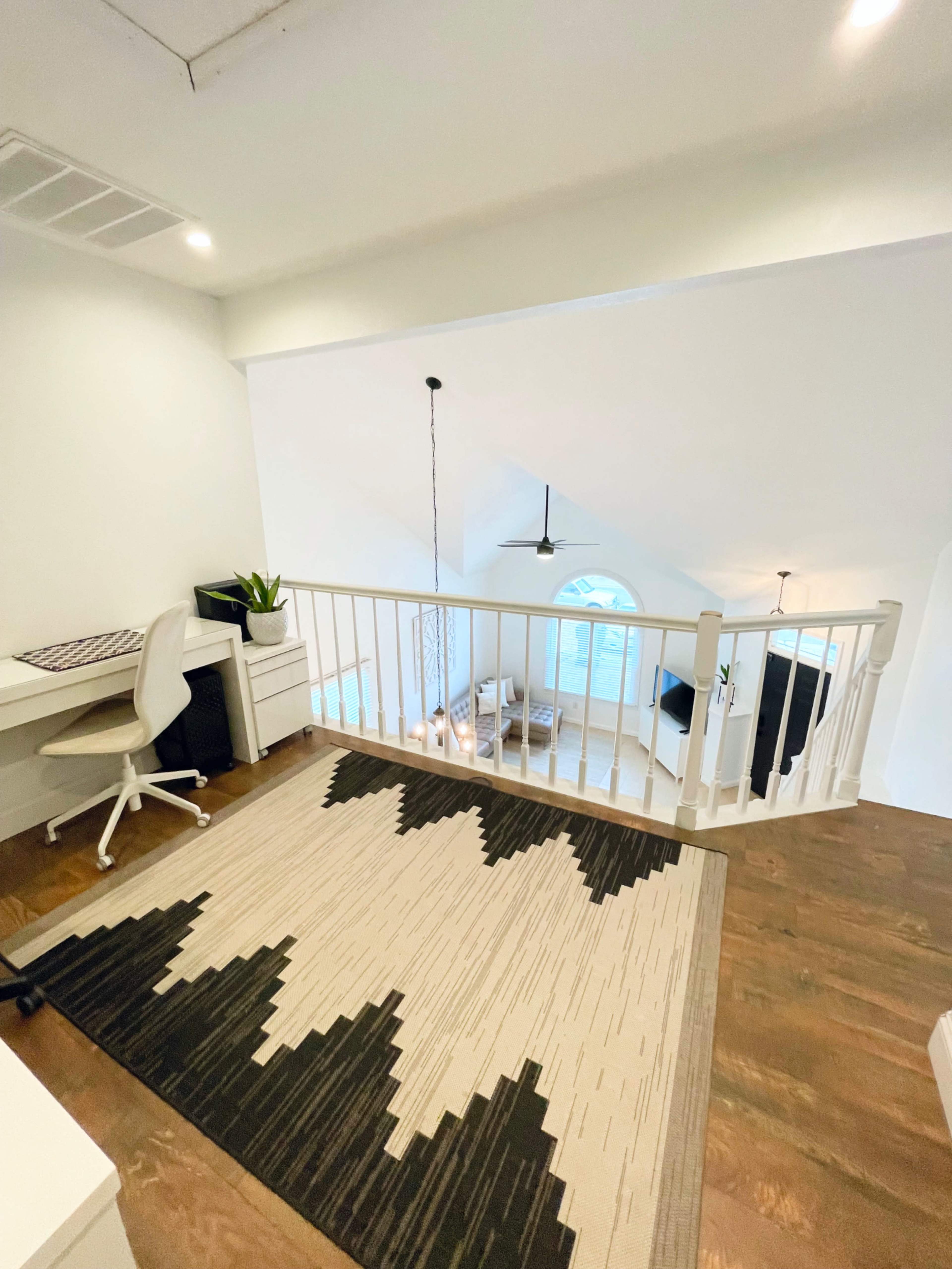 A well-lit loft space features a desk and chair near a white railing, with a geometric rug on the wooden floor and a large window providing natural light.