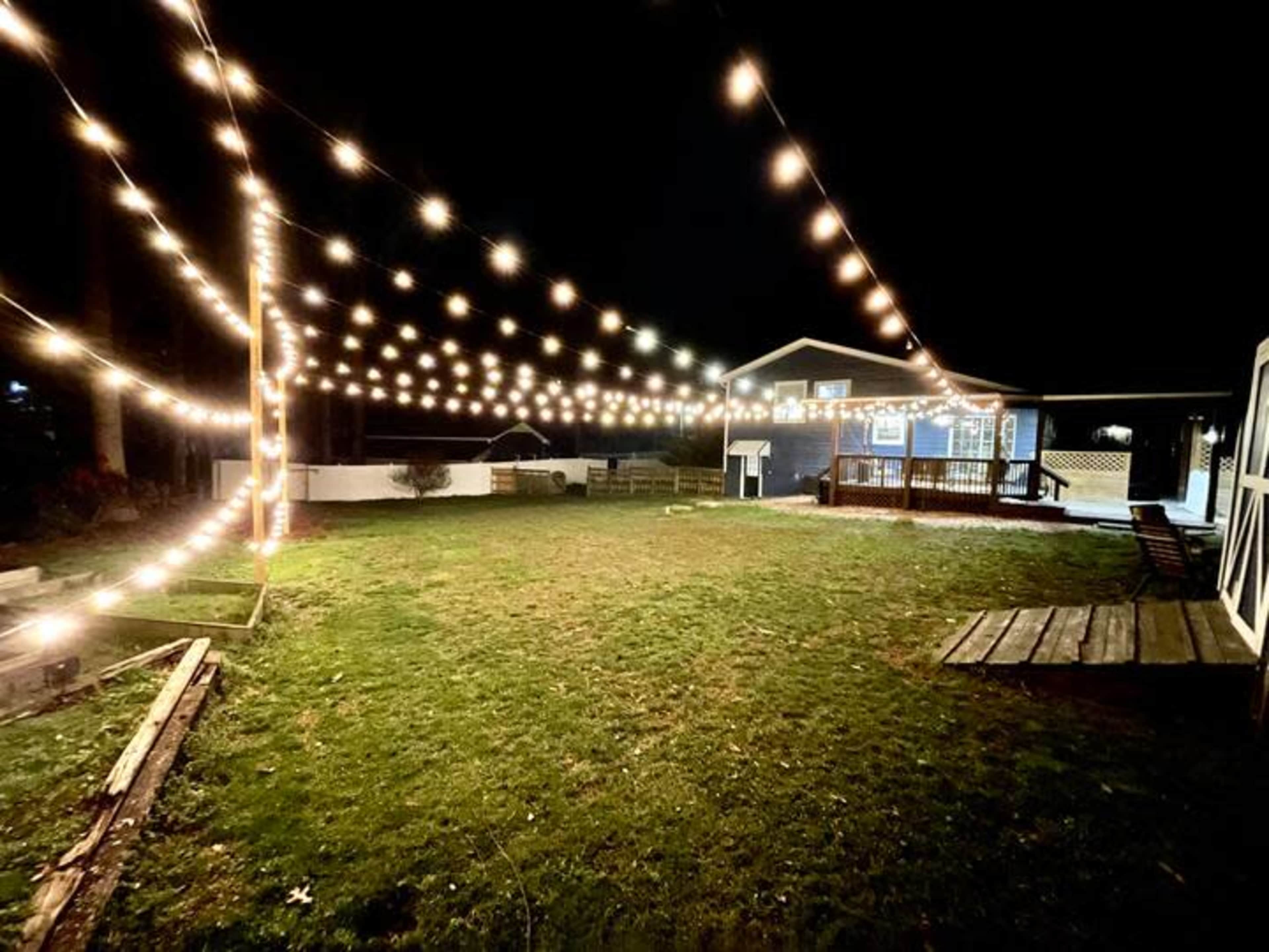 A backyard is illuminated by string lights, with a wooden deck and a house visible in the background at night.