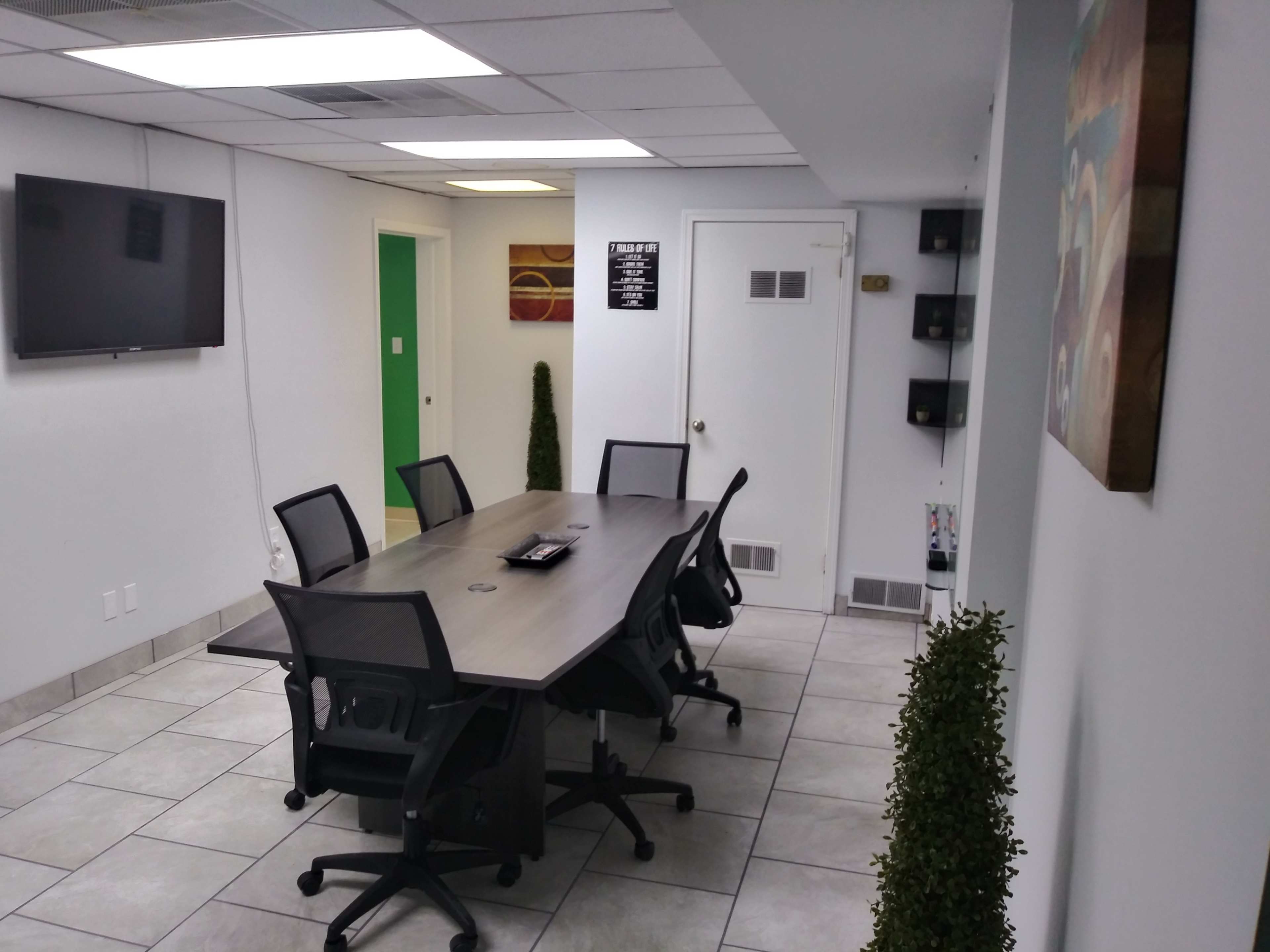 A conference room with a rectangular table surrounded by rolling office chairs, a television mounted on the wall, and potted plants in the corners.