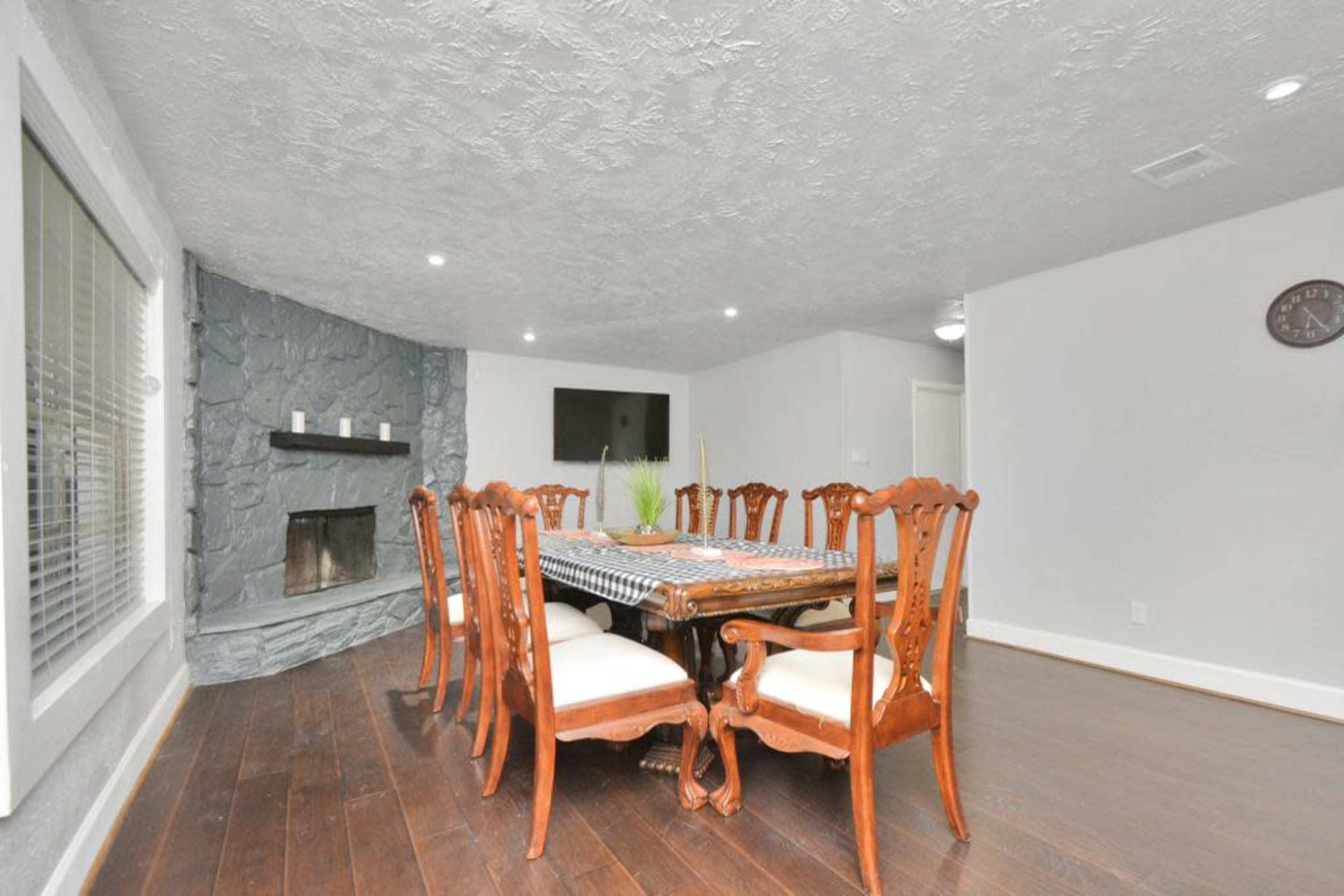 A dining room features a large wooden table surrounded by ornate chairs, with a stone fireplace and a TV mounted on the wall.