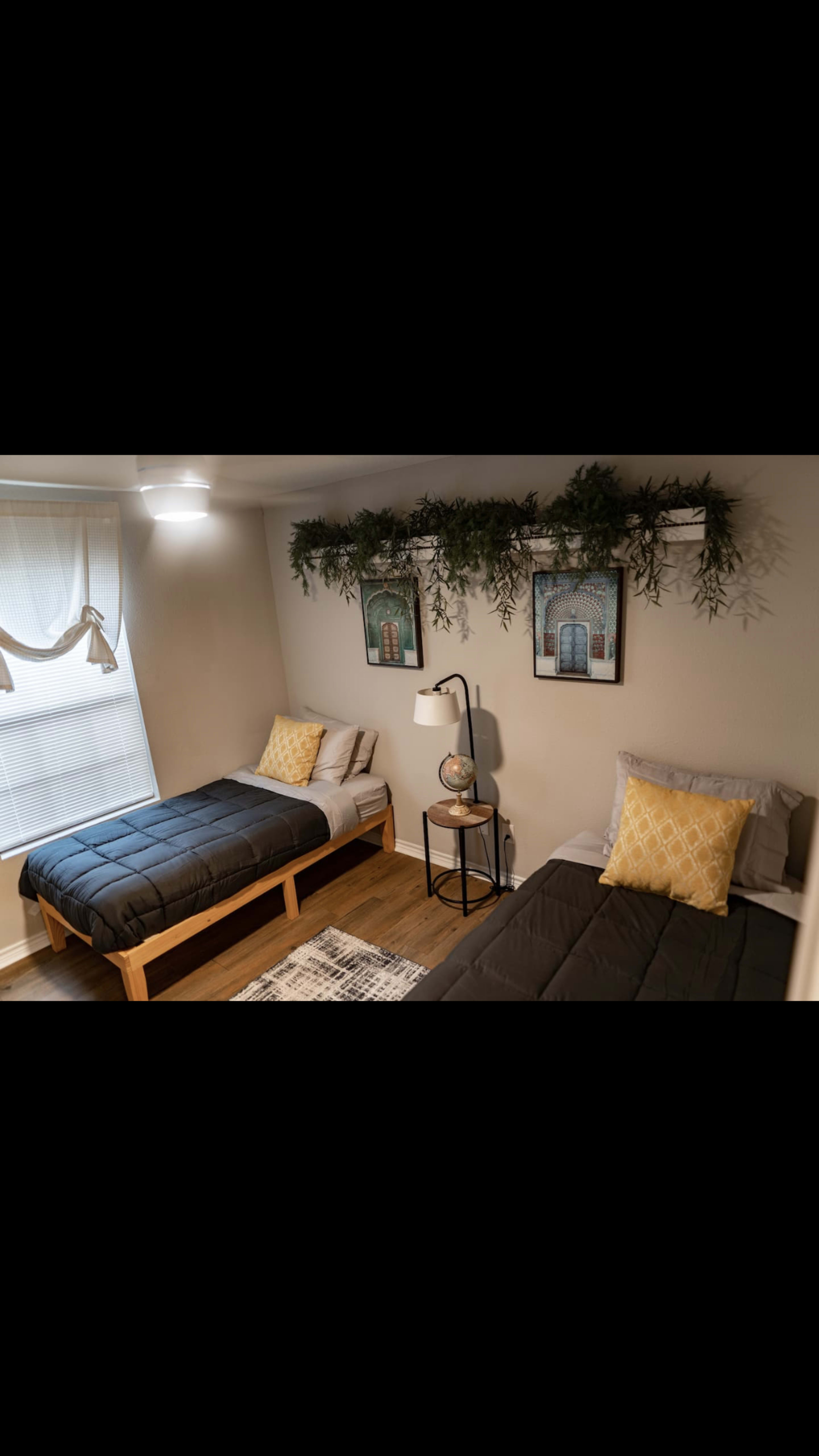 A small bedroom featuring two twin beds, decorative wall art, and plants arranged above a nightstand with a lamp and a globe.
