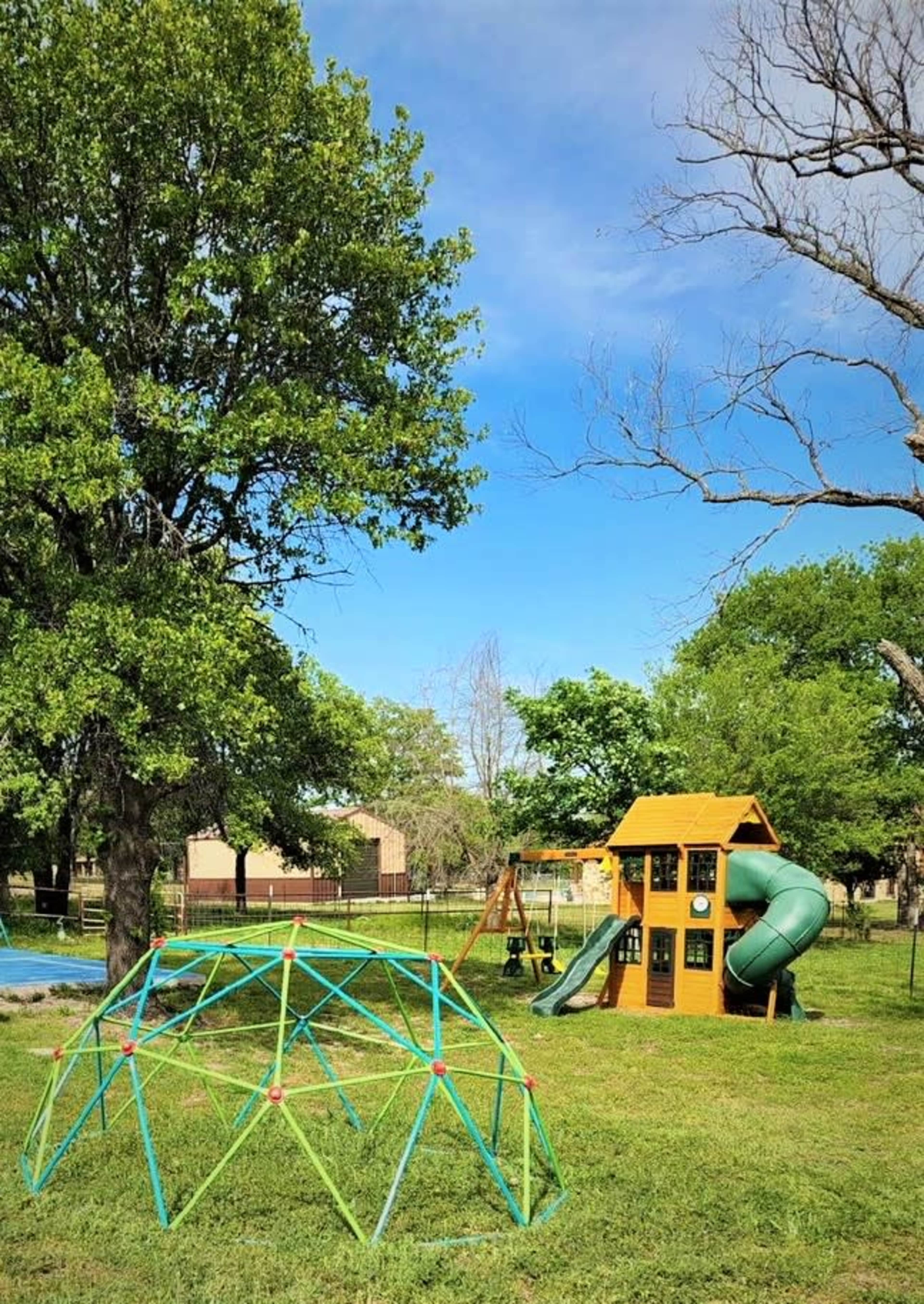 The scene depicts a playground featuring a colorful climbing dome and a playset with a slide, set in a grassy area with a few trees under a clear blue sky.