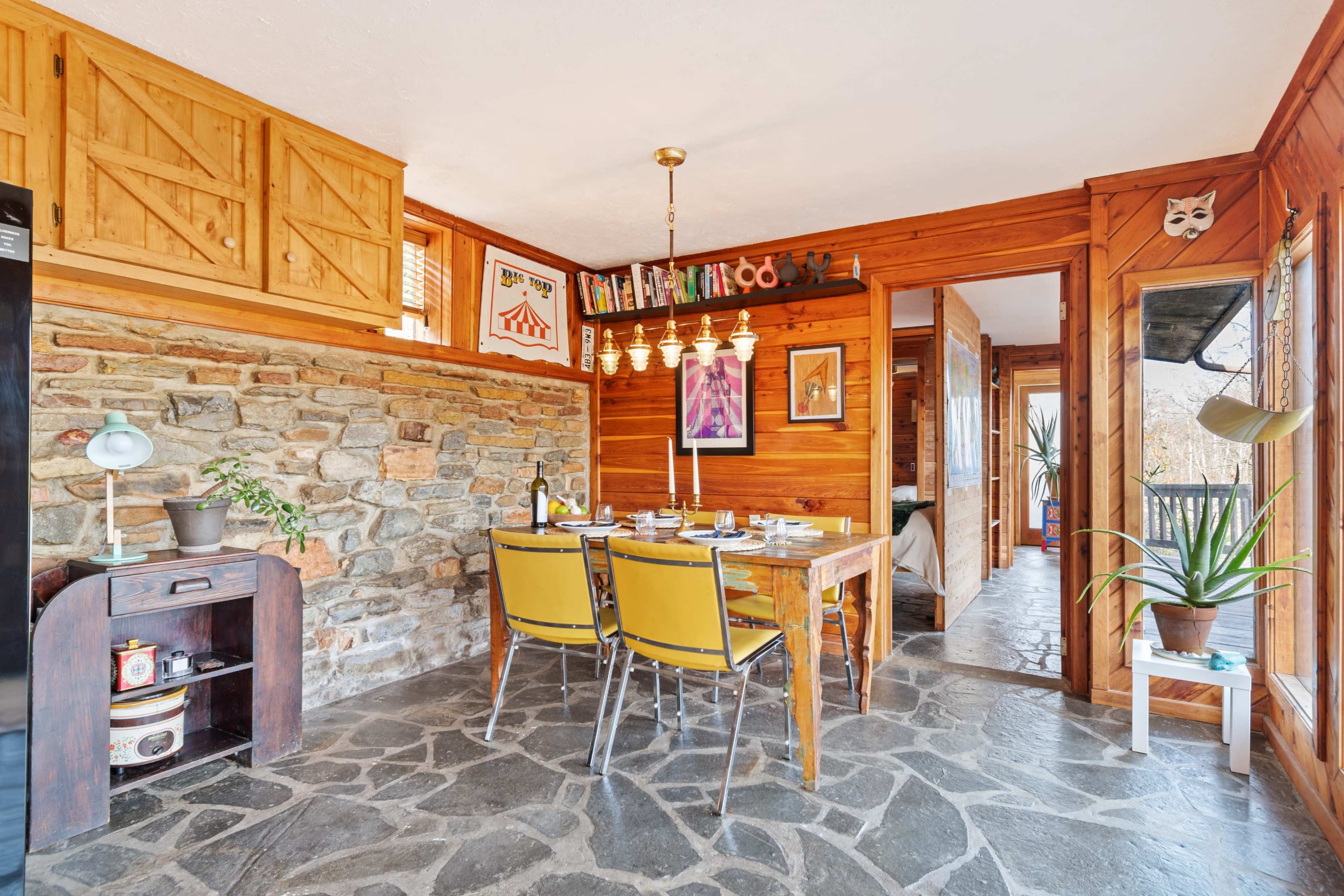 A dining area with a wooden table surrounded by yellow chairs, stone walls, and framed artwork, leading to a doorway and another room.