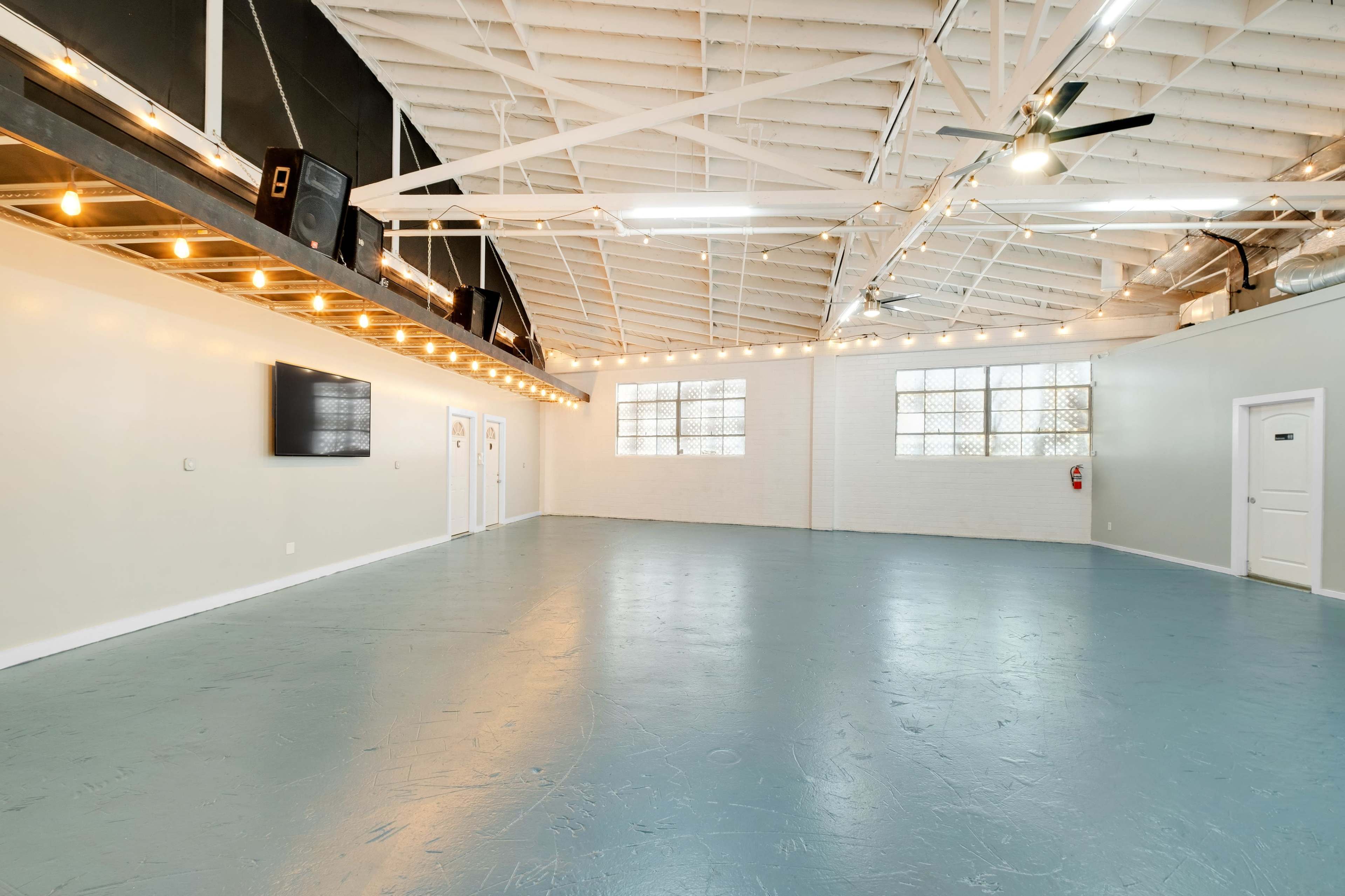 A spacious, empty room with high ceilings, large windows, and a polished blue floor, illuminated by string lights.