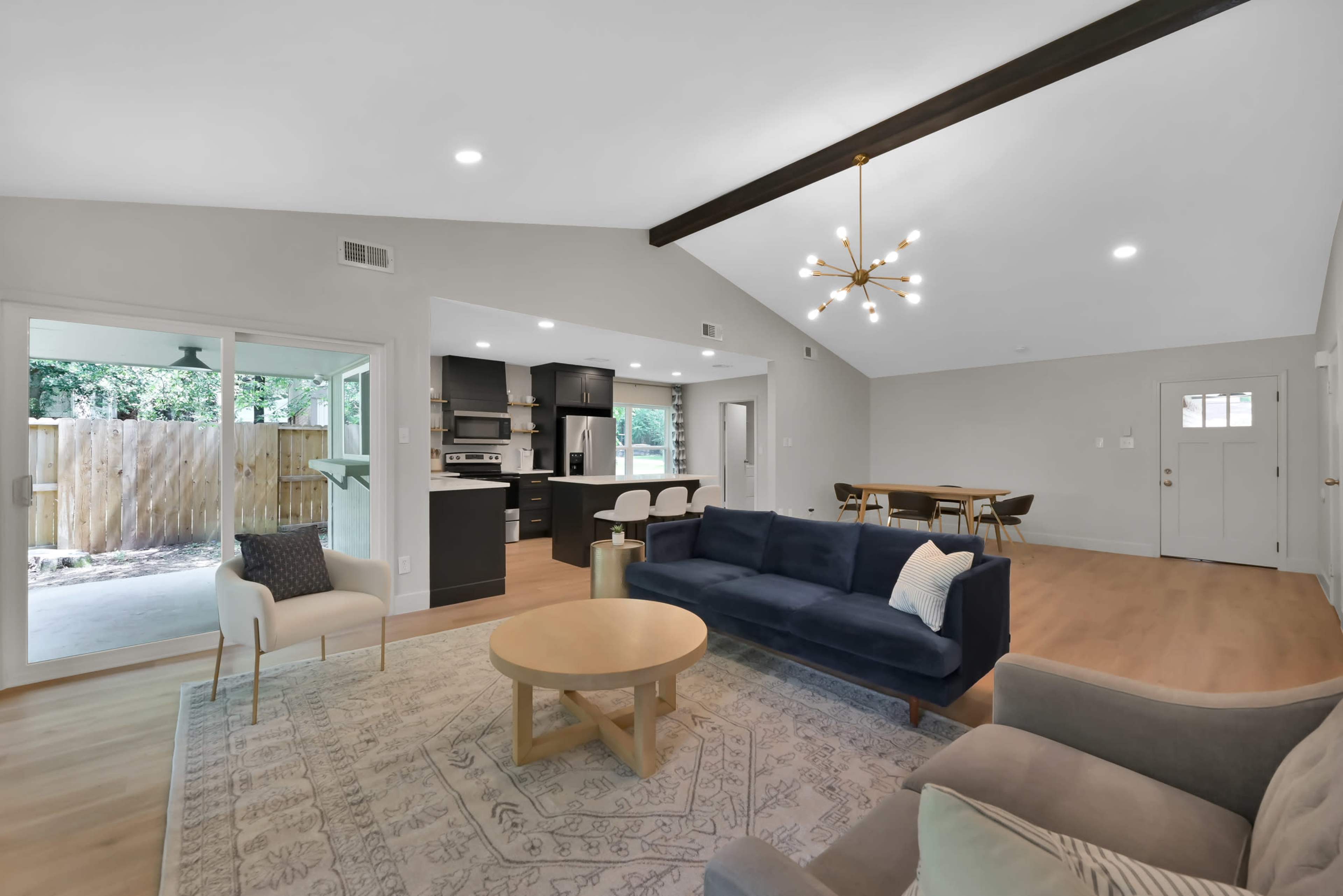 A modern living room with a blue sofa, a round wooden coffee table, and a dining area in the background, featuring a kitchen with black cabinetry.
