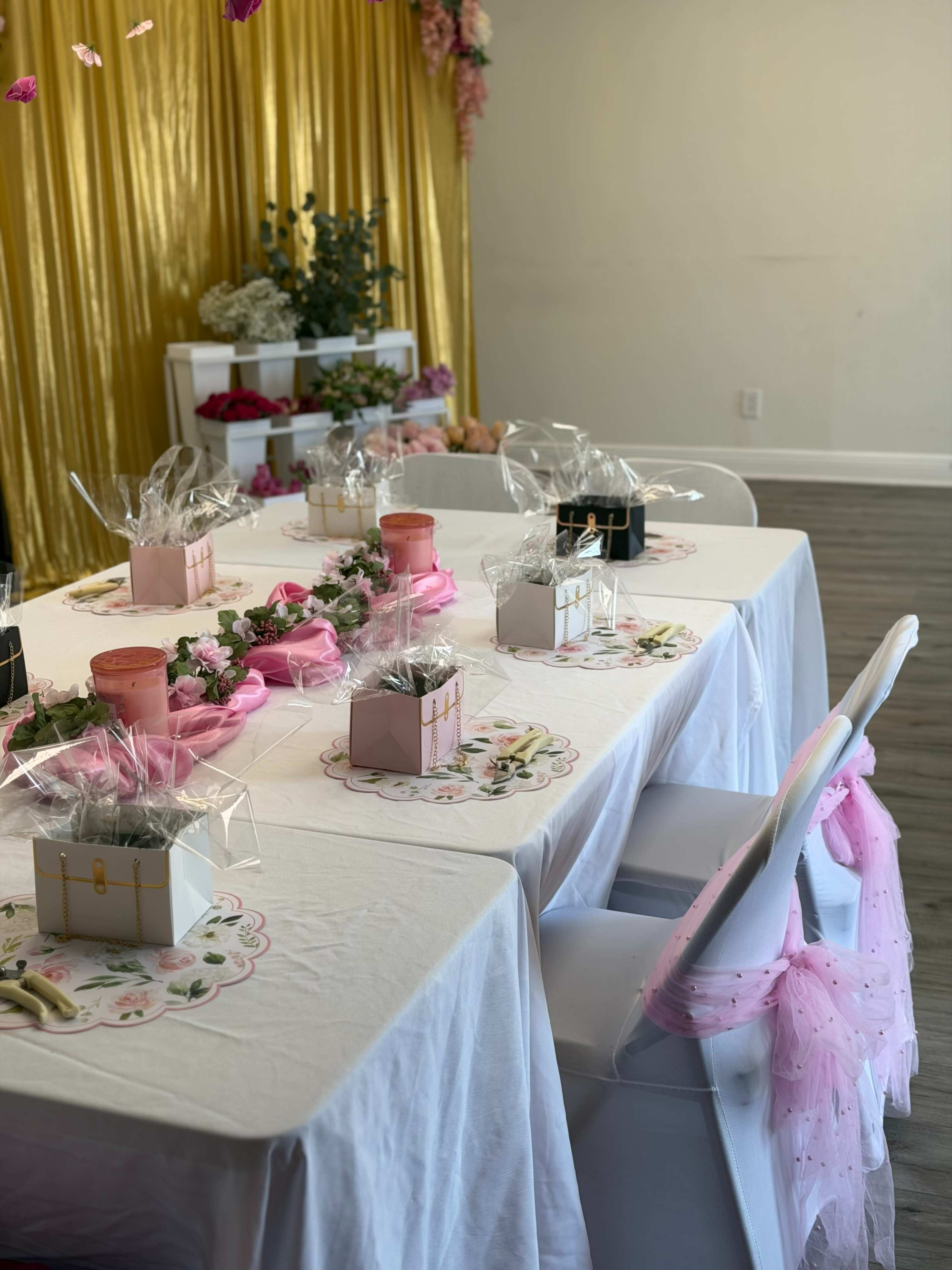 A decorated table is set with white tablecloths, pink gift boxes, floral centerpieces, and a backdrop of gold drapes.