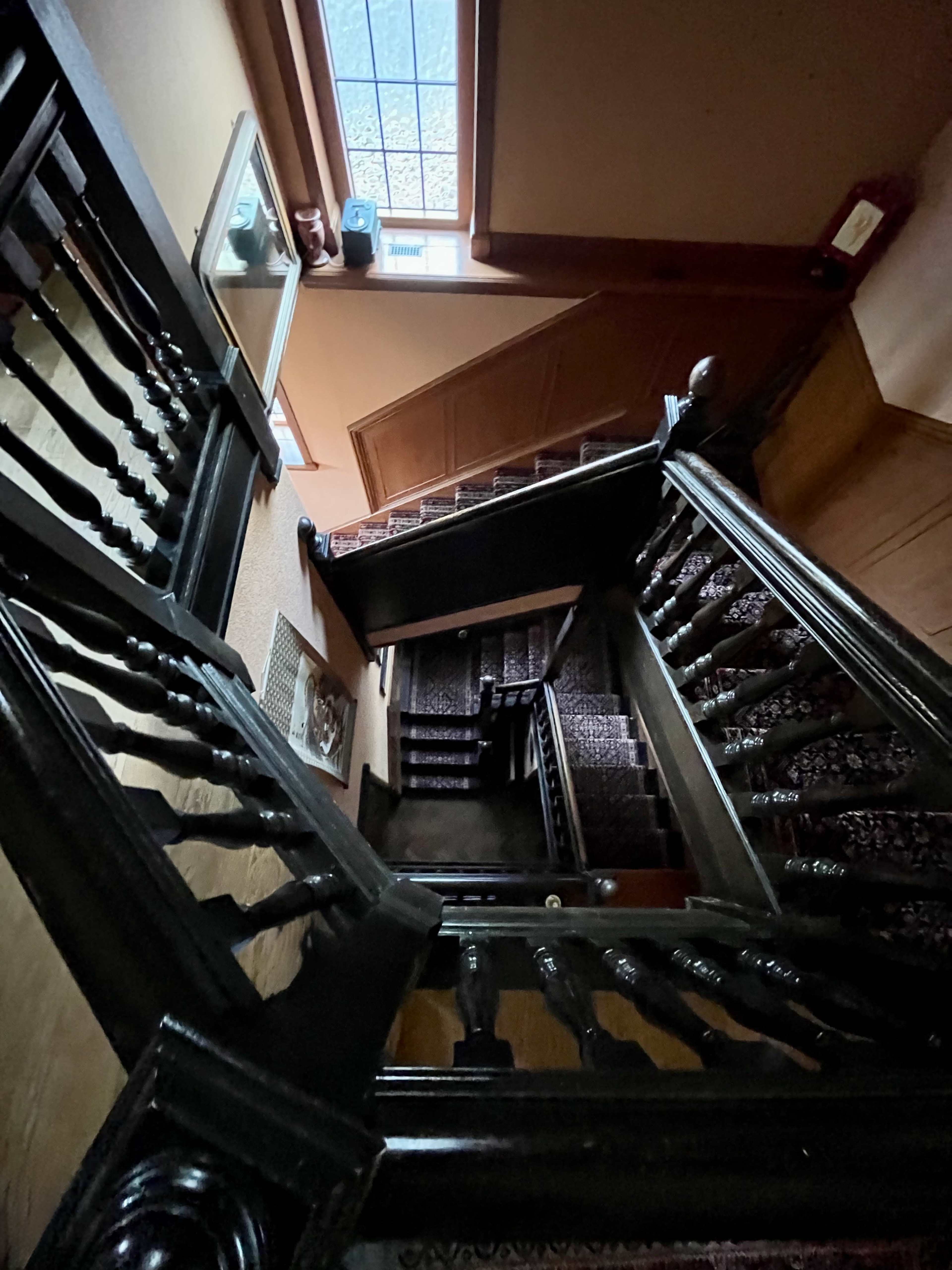 The image shows a top-down view of a wooden staircase with a patterned carpet, leading downward in a well-lit hallway.