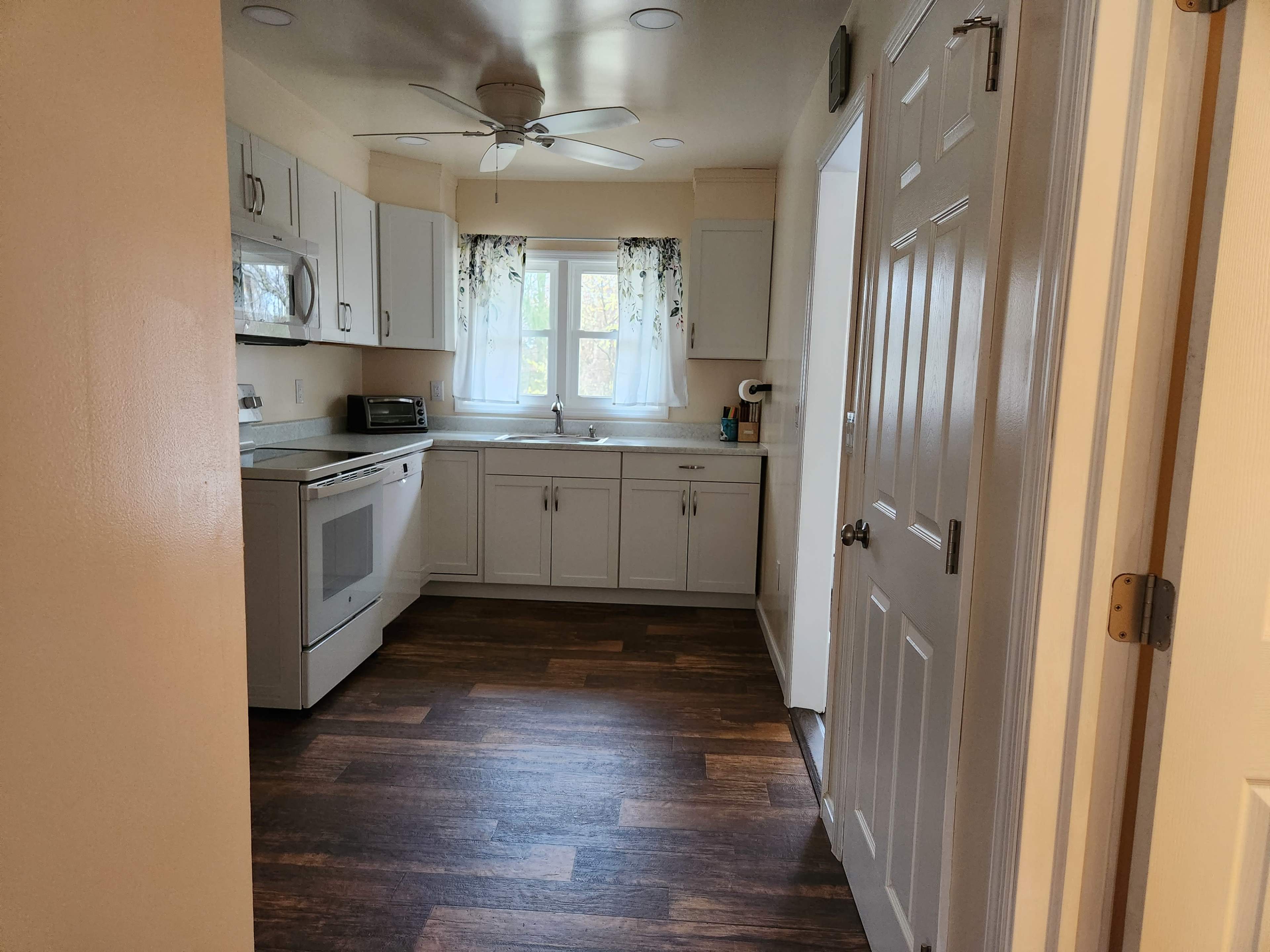 The image shows a well-lit kitchen featuring white cabinetry, a window with sheer curtains, and a door leading to another room.