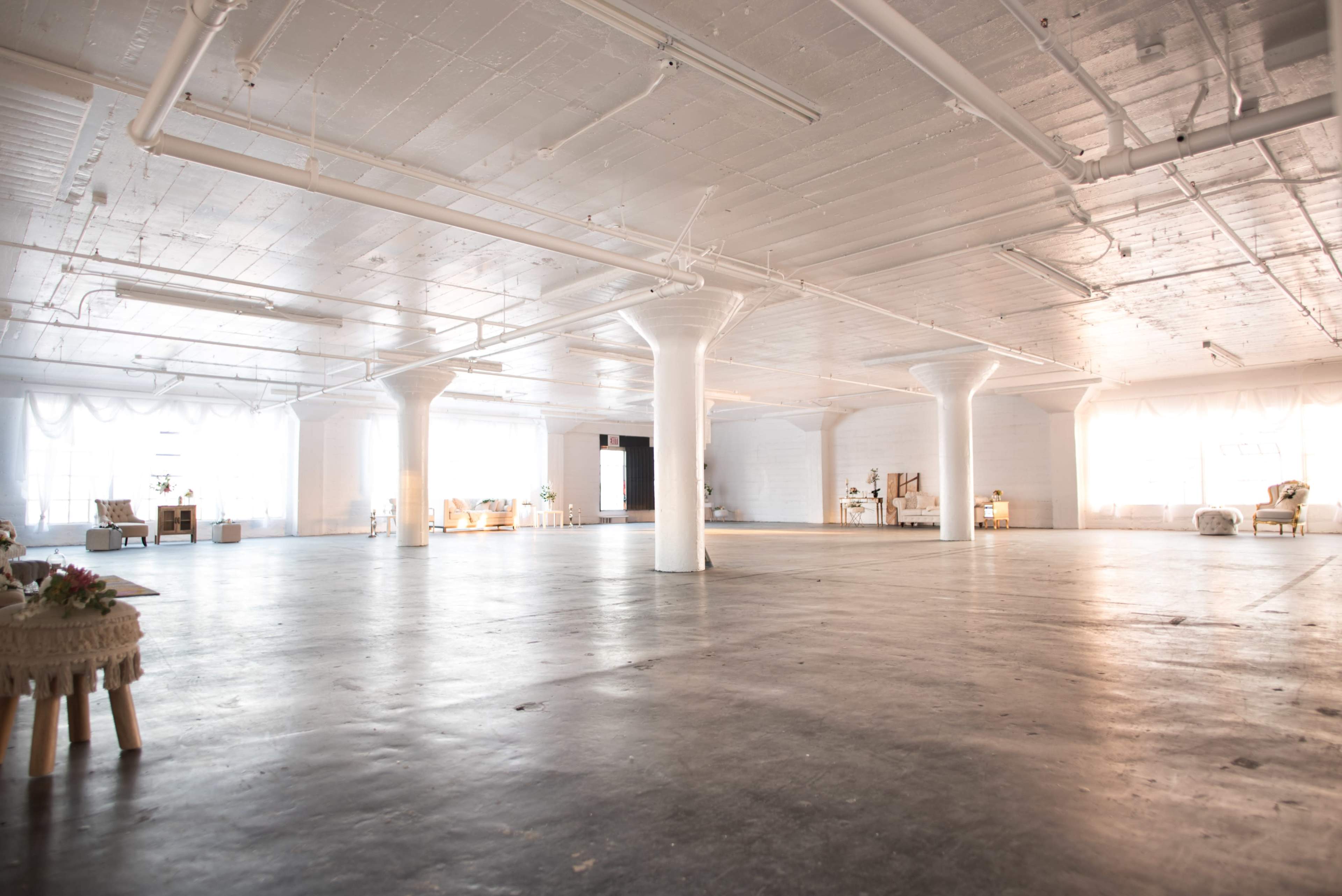 A large, empty industrial space features white walls, polished concrete floors, and several columns, with natural light streaming in from large windows.