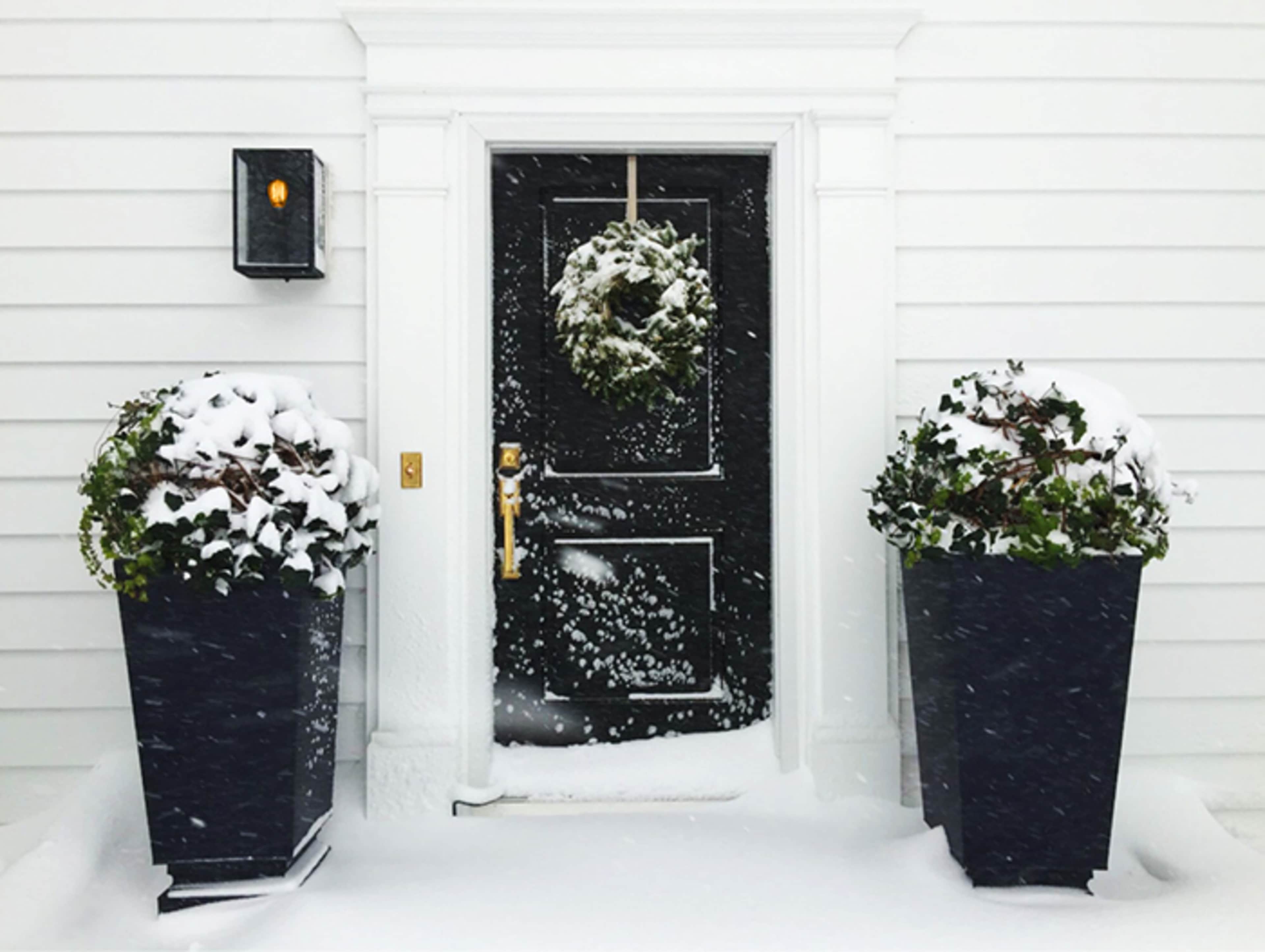 A black front door with a wreath is flanked by two large planters covered in snow, with drifted snow on the ground in front of the door.