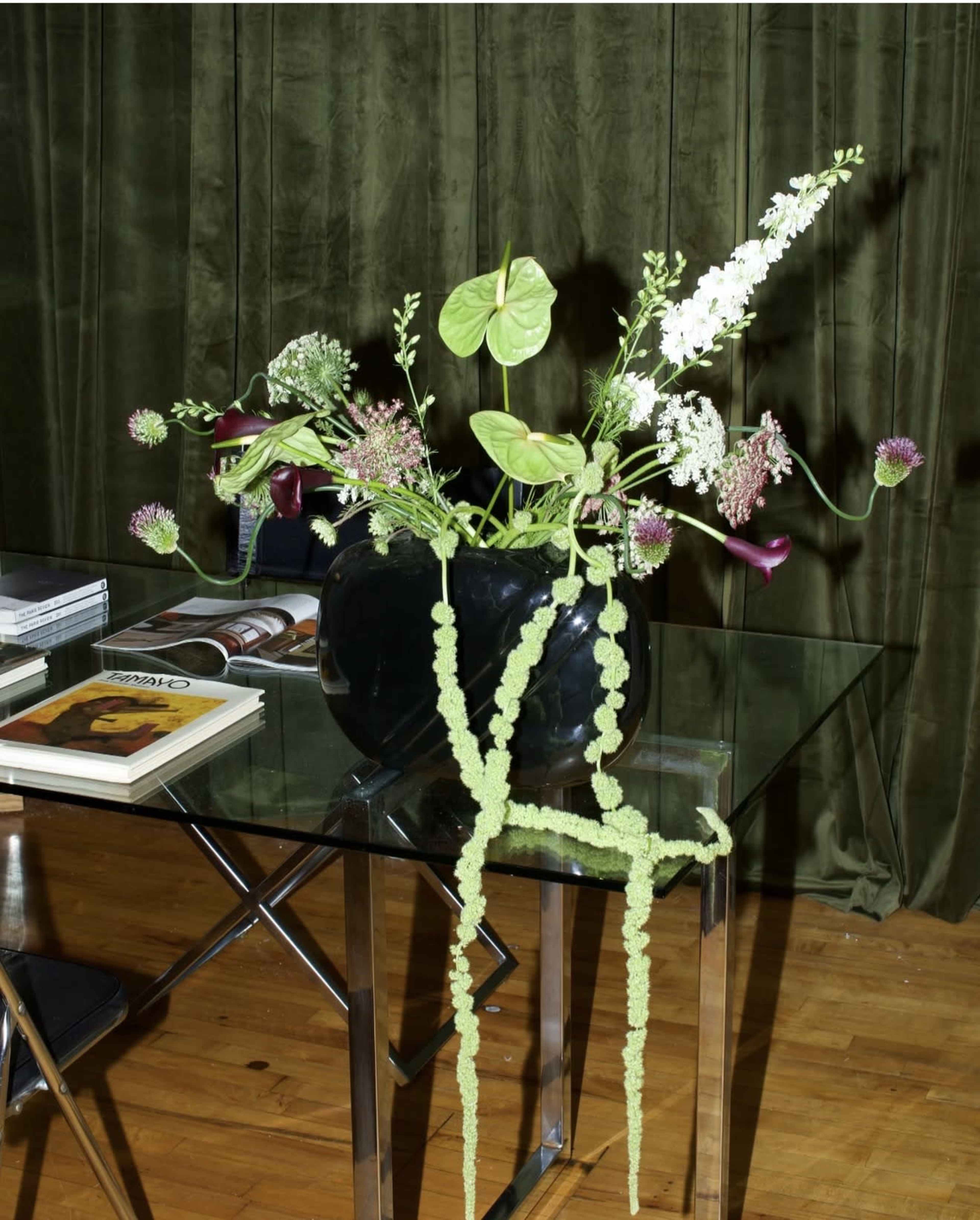 A black vase filled with a variety of flowers and greenery sits on a glass table against a green fabric backdrop.