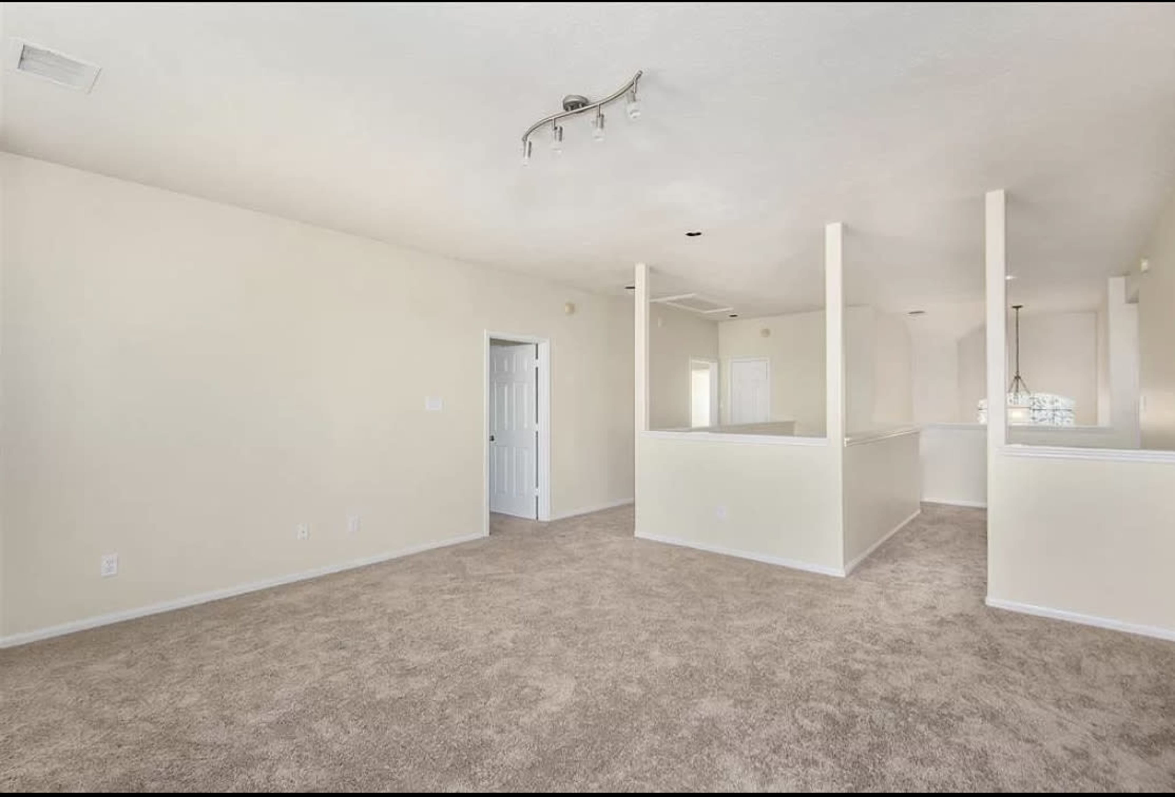 An empty living room with beige carpet and light-colored walls, featuring a partial divider leading to an open kitchen area.