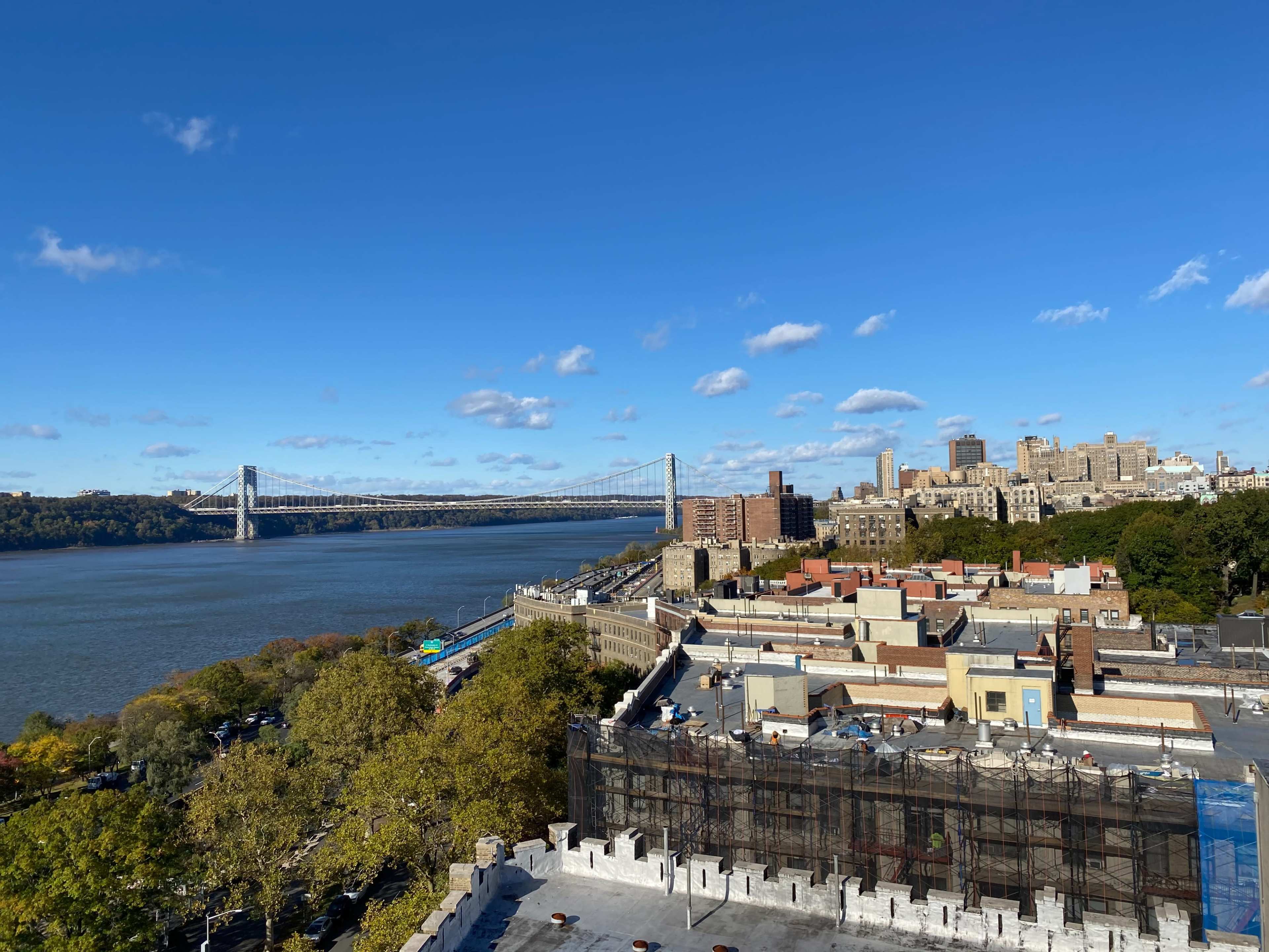 The image shows a view of the Hudson River with the George Washington Bridge in the background, and buildings lining the riverbank, including a neighborhood with low-rise structures and trees.
