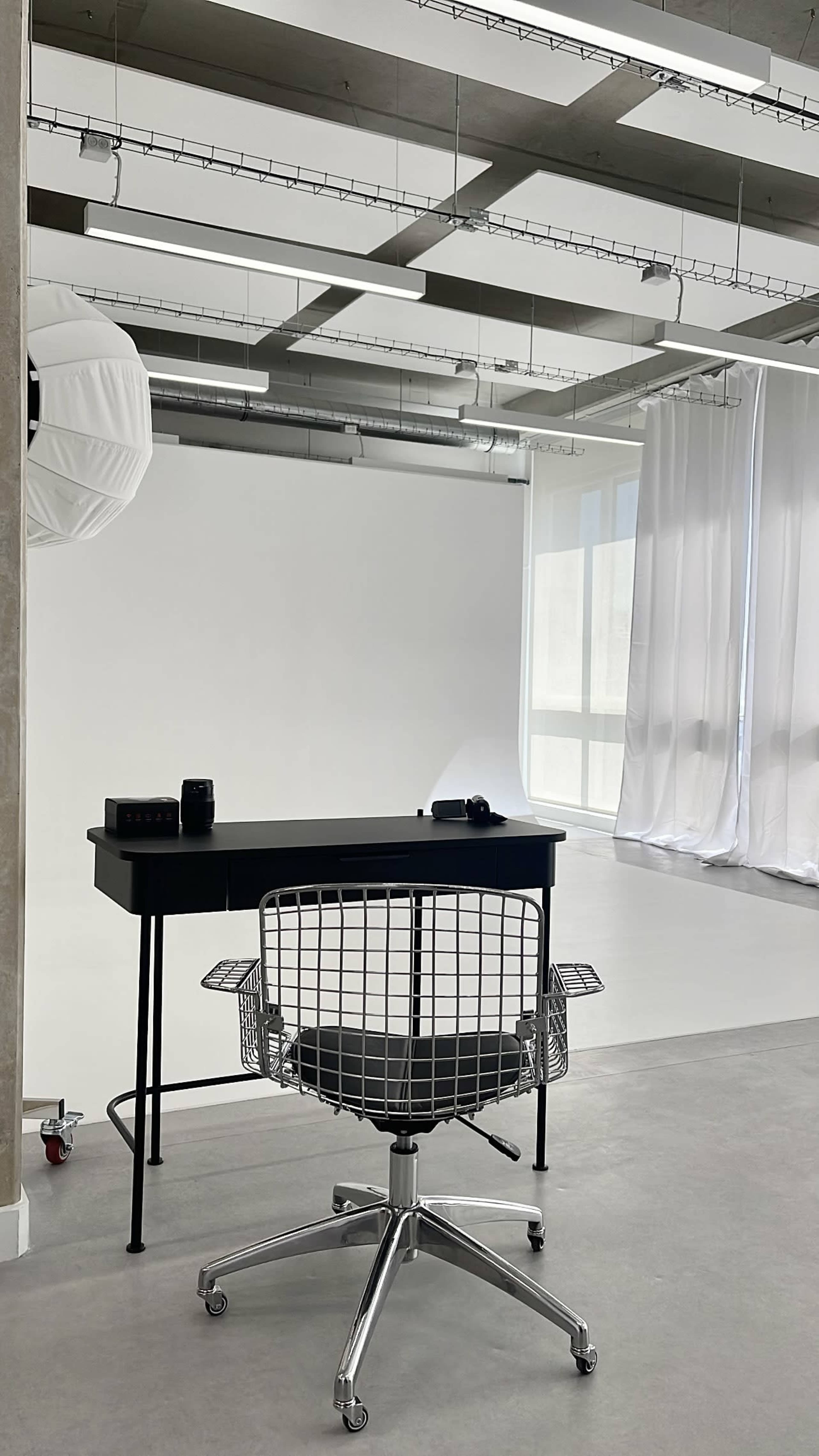 A minimalist office space features a black desk with a chair on wheels, set against a backdrop of white curtains and a smooth floor.