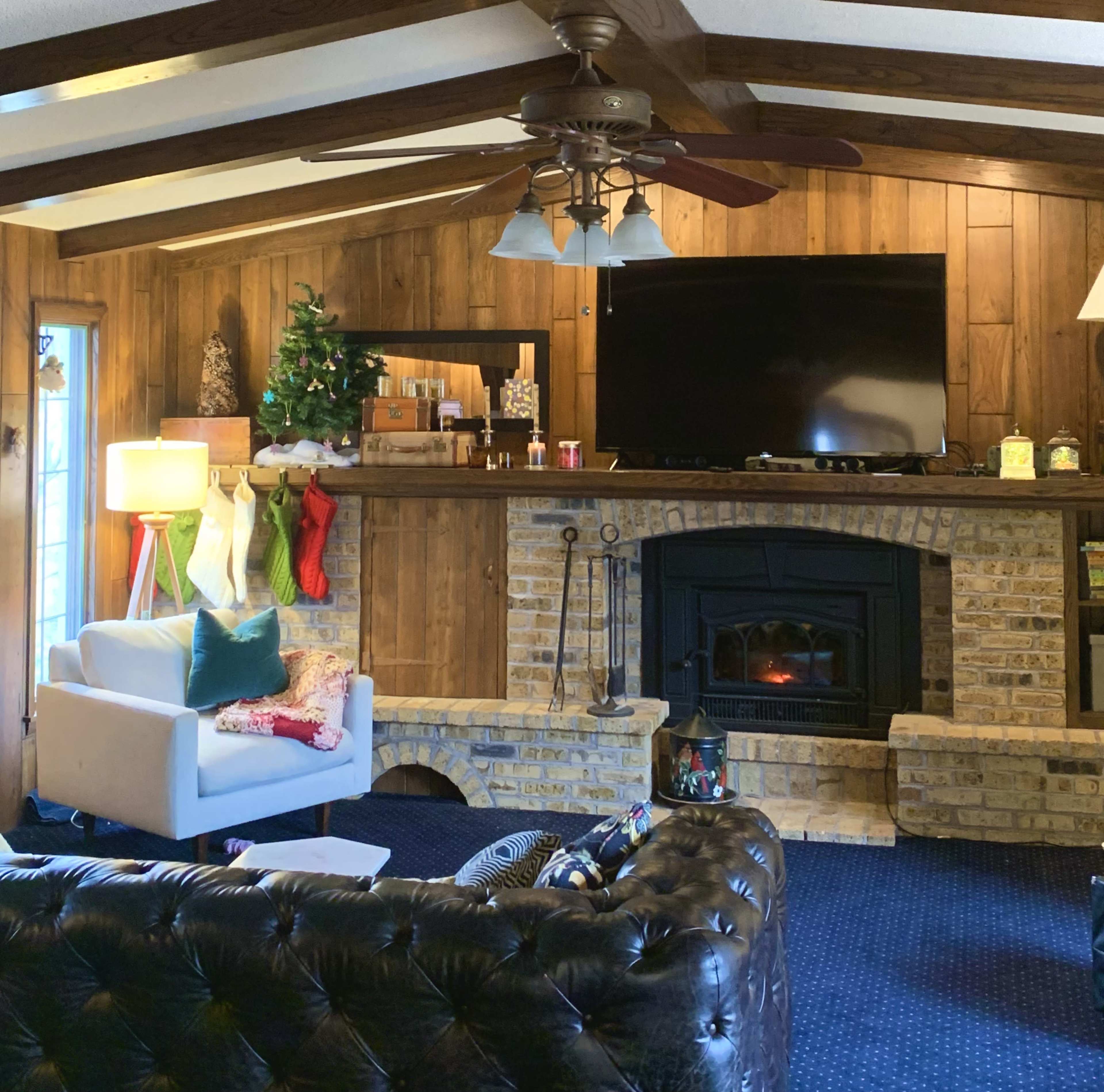 A cozy living room with wooden walls, a stone fireplace, a large television above the mantel, and festive decorations, including a small Christmas tree and stockings.