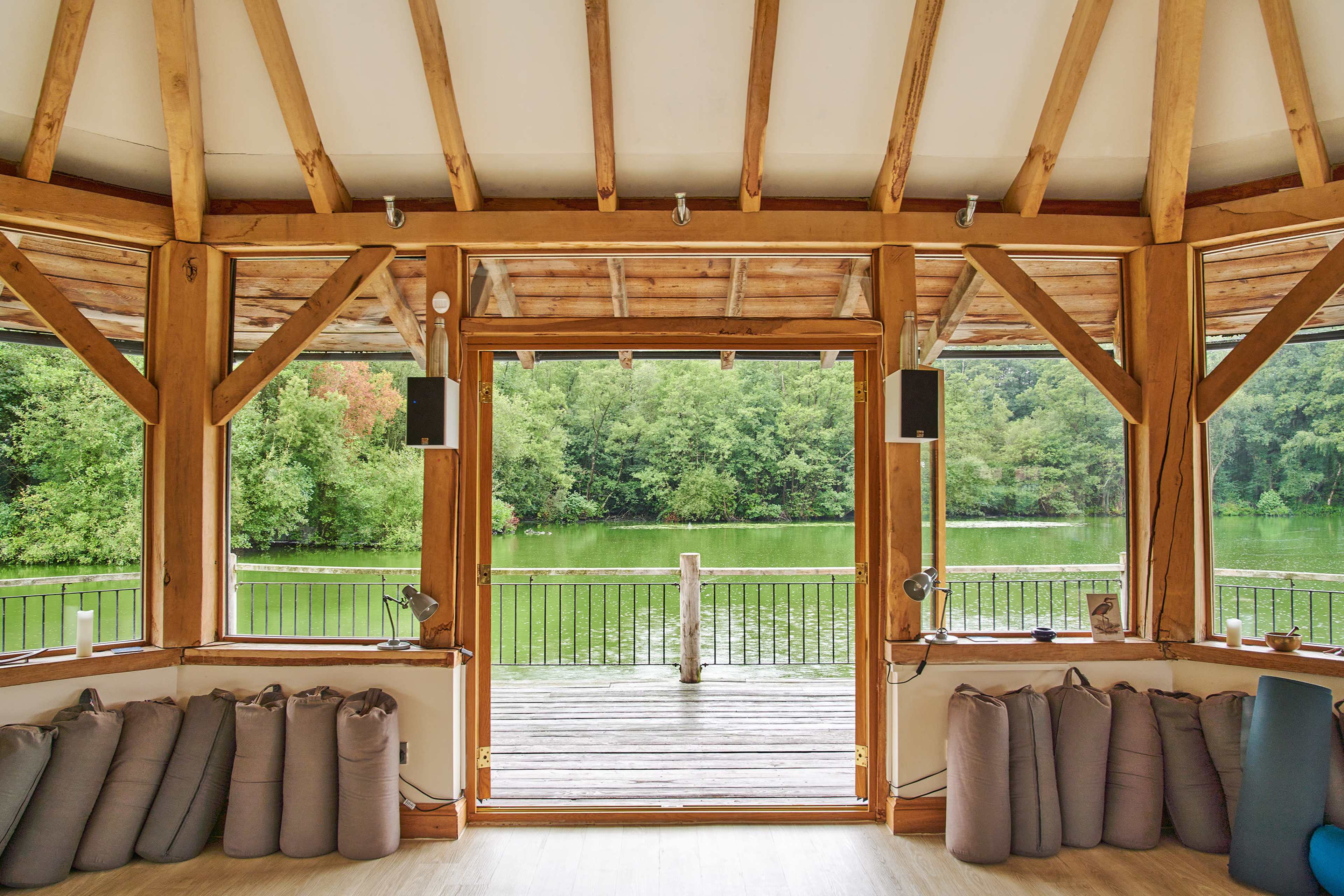 The image shows a wooden interior with large windows facing a green lake surrounded by trees, and seating cushions neatly arranged along the walls.