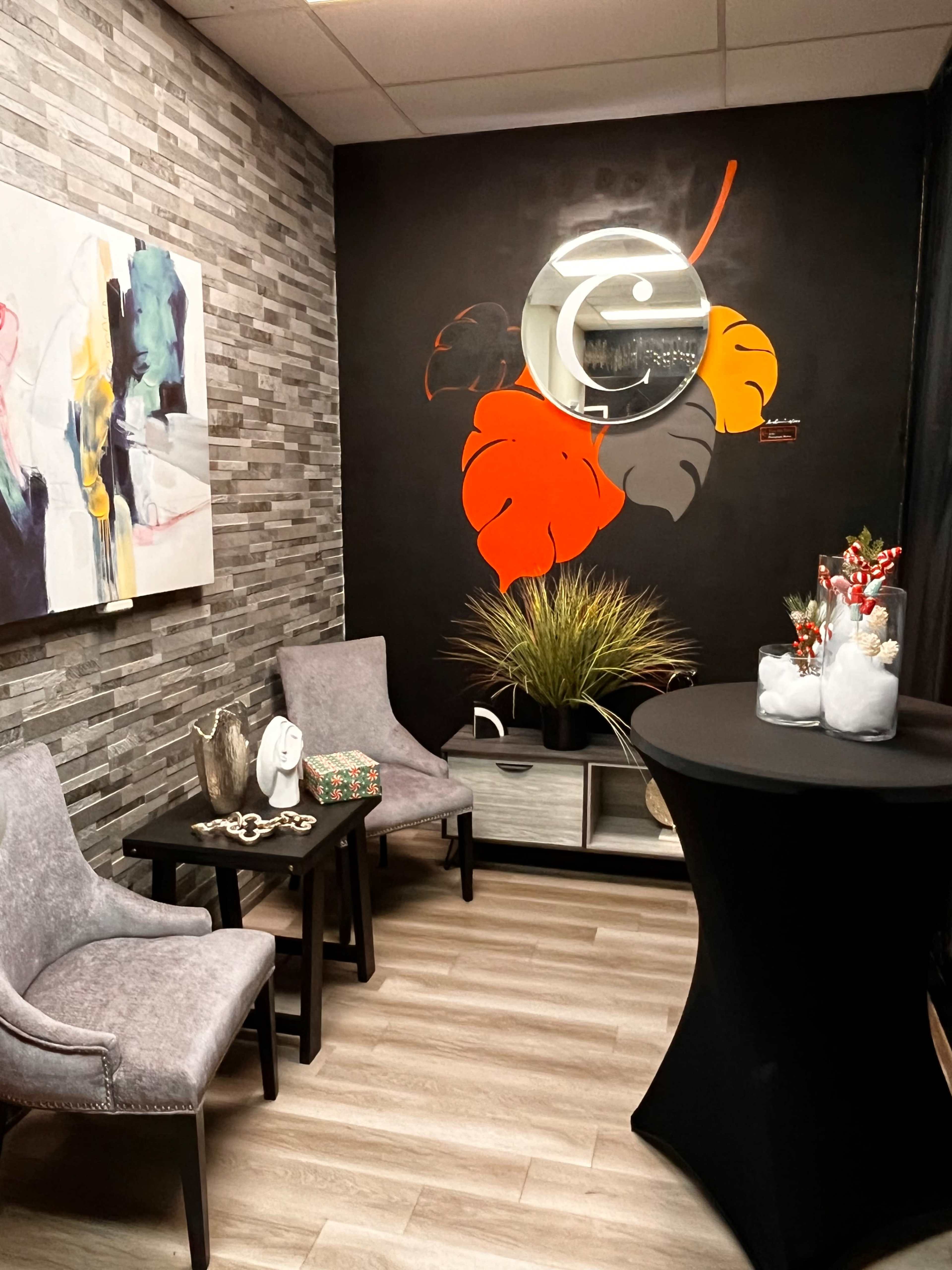 The image shows a cozy waiting area featuring two gray chairs, a small black table, and decorative elements such as artwork and indoor plants against a black accent wall.