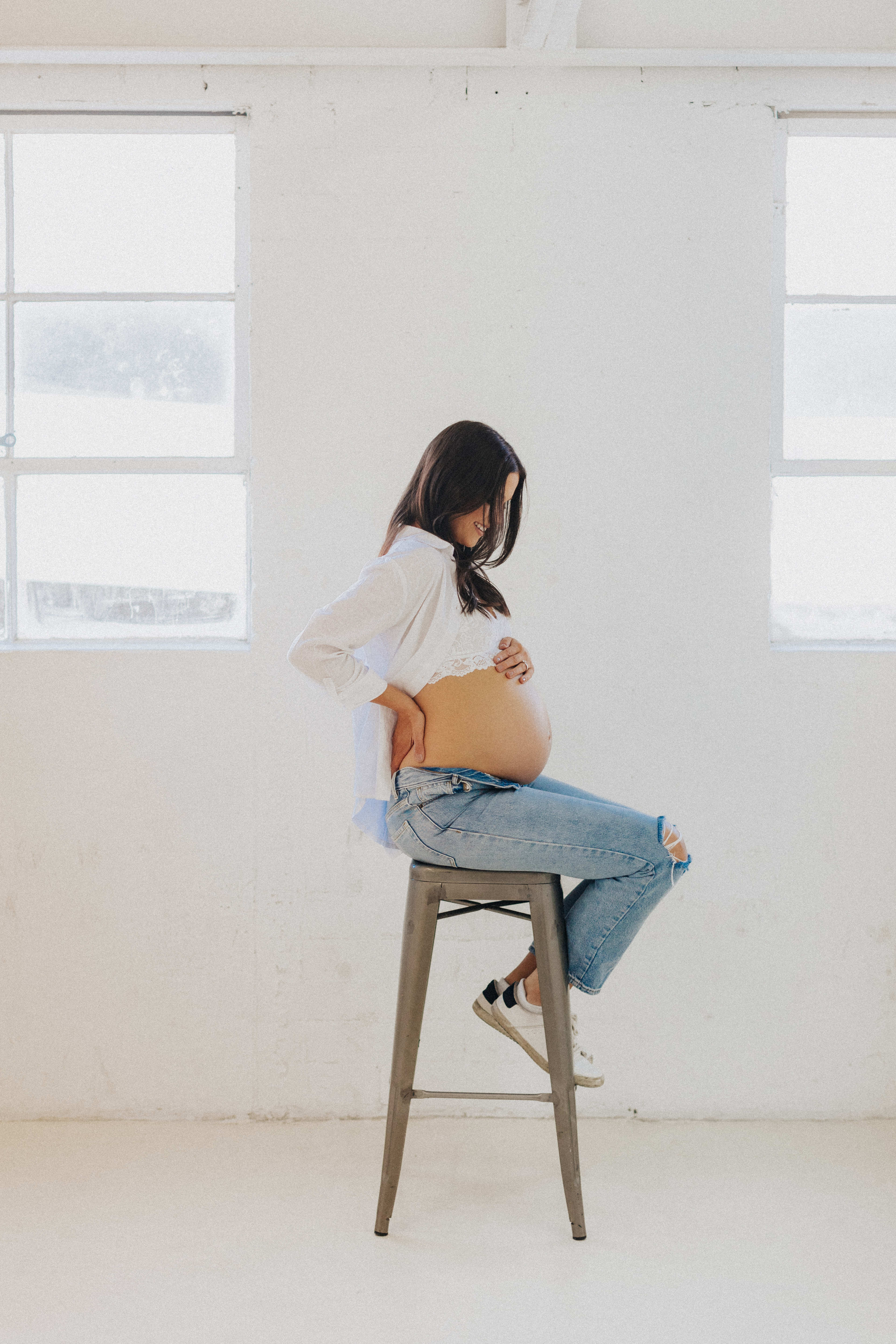 Una sesión de fotos de una mujer embarazada en un taburete blanco con un decorado minimalista.