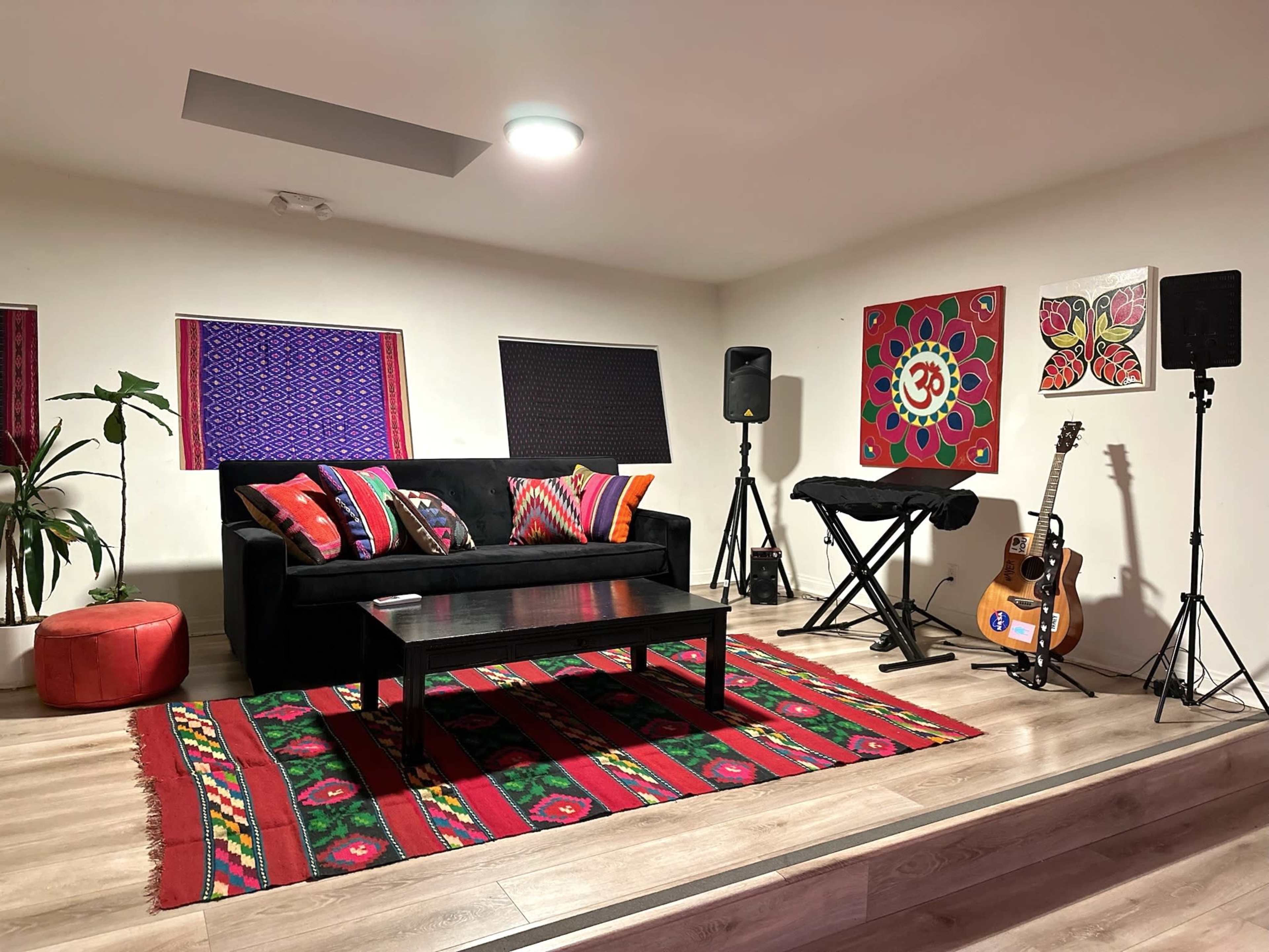 The room features a black couch with colorful cushions, a wooden coffee table, a decorative rug, and musical equipment, including a guitar and keyboard.
