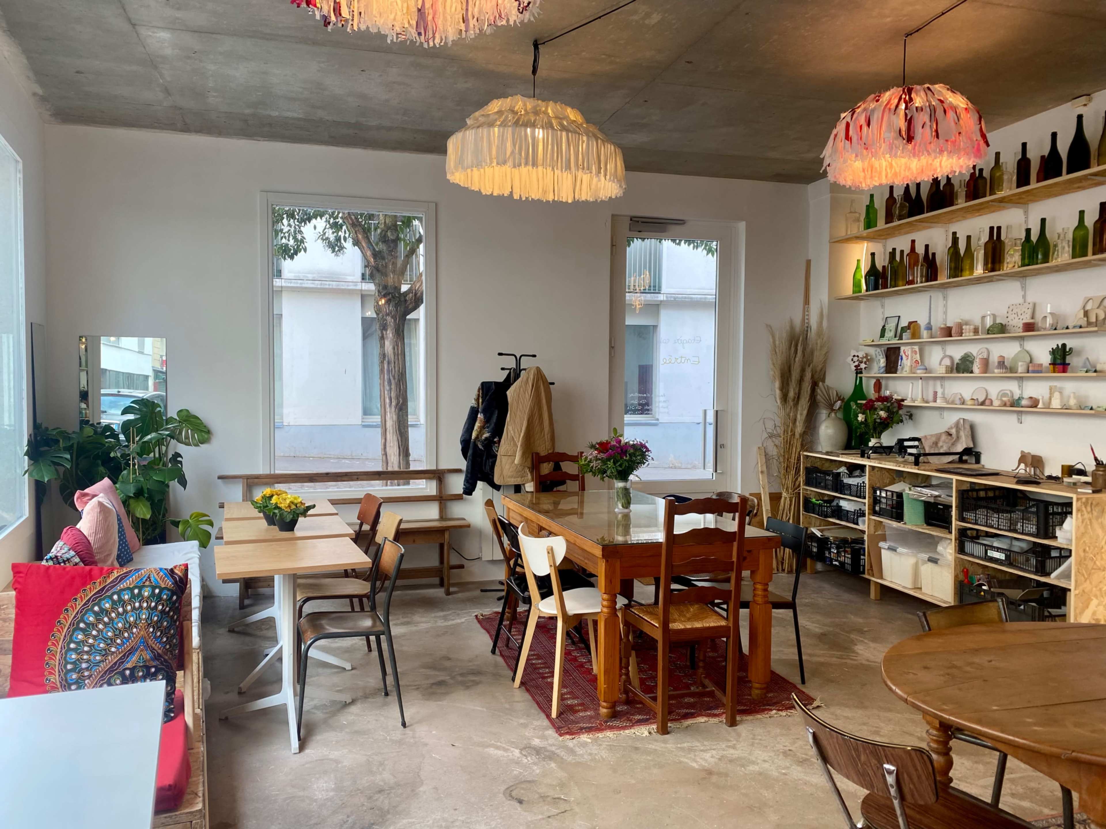 A cozy interior space features wooden tables, chairs, and plants, with a wall shelf displaying bottles and decor.