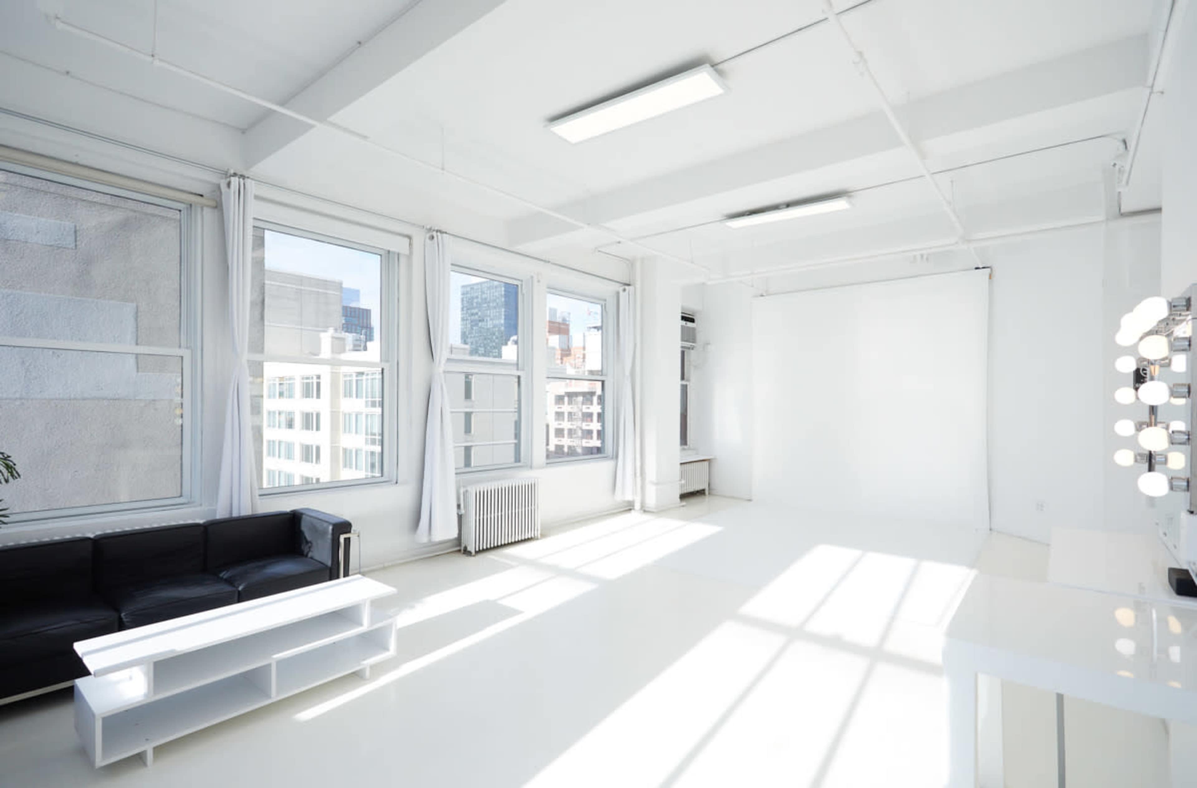 The image shows a bright and open studio space with large windows, a black couch, and a white backdrop area.