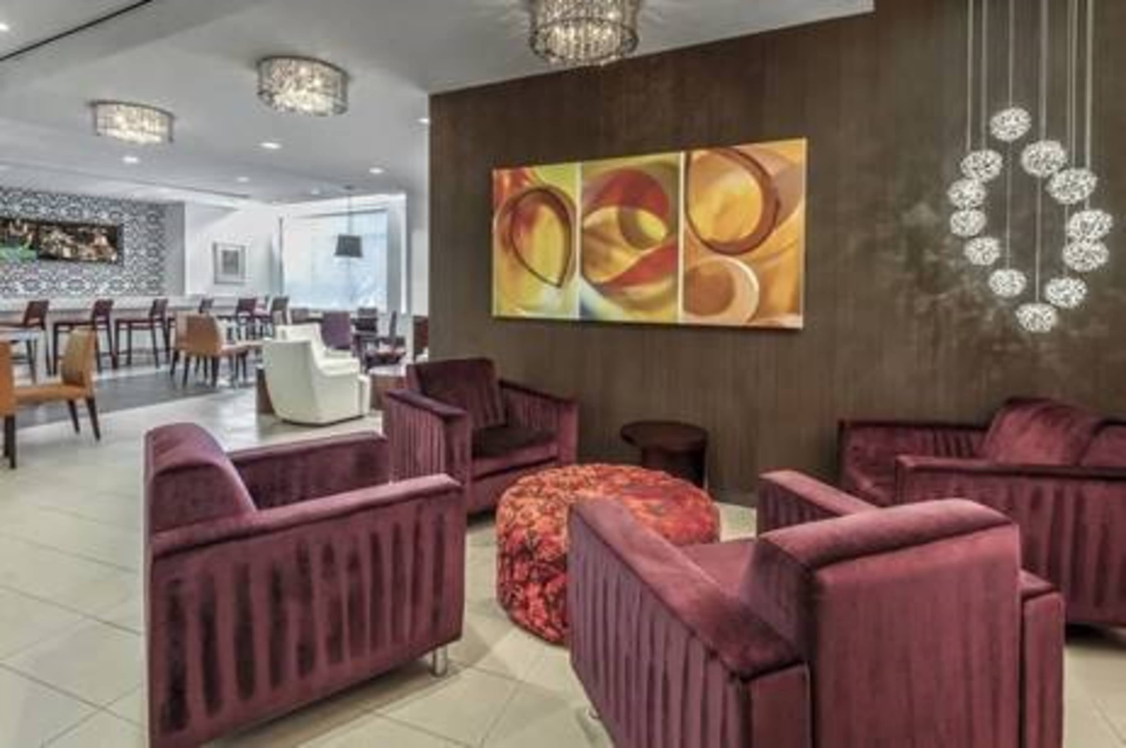 A modern lounge area featuring sleek purple armchairs, a round patterned ottoman, and decorative wall art under stylish light fixtures.