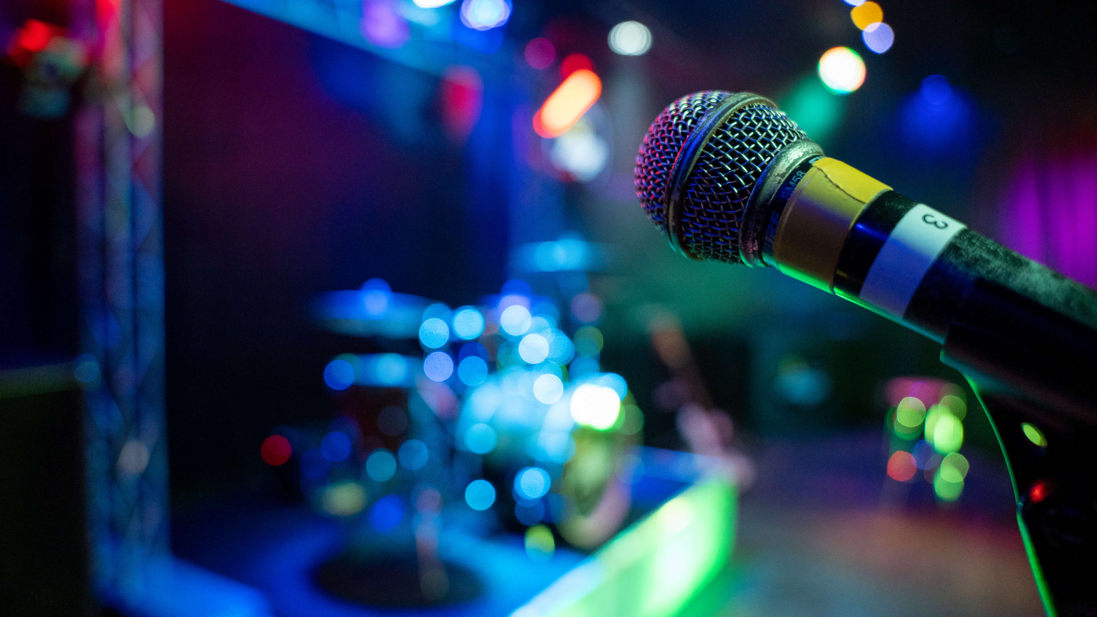 A microphone stands in the foreground, with a blurred drum set and colorful stage lights illuminated in the background.