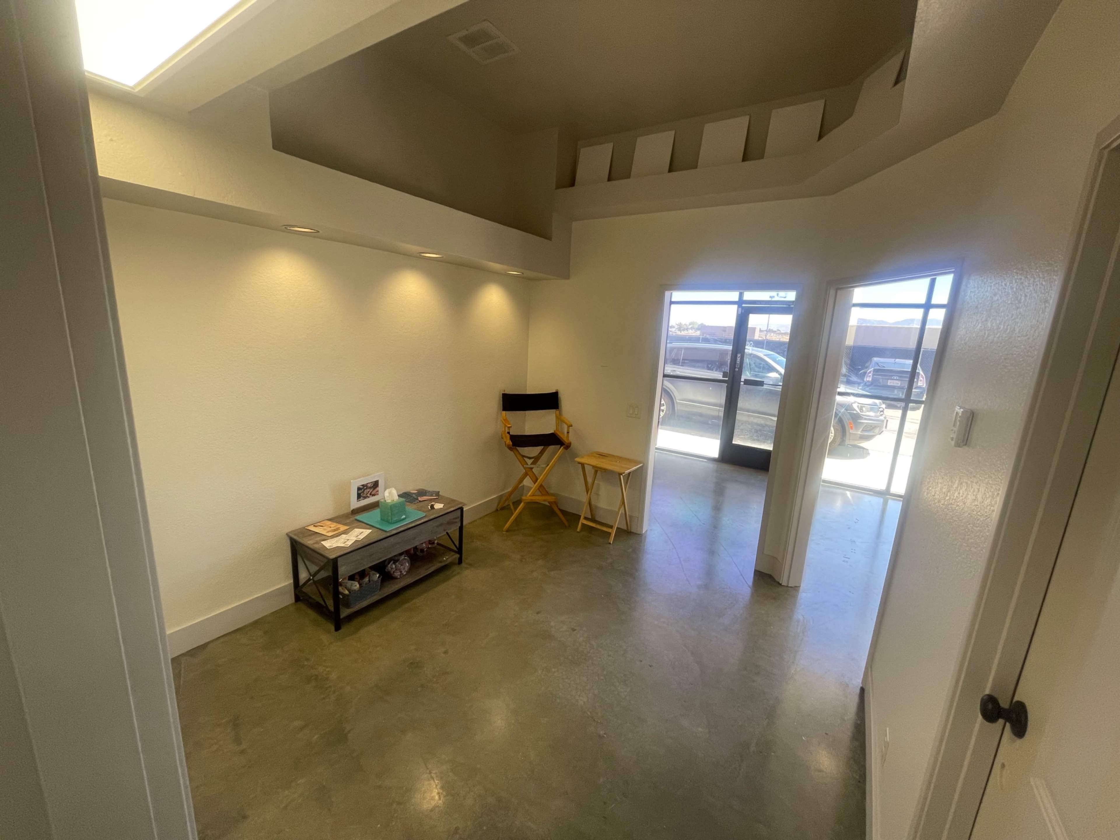 The image shows a bright, minimalistic interior space featuring a small table with magazines and a director's chair, along with a large window looking out to the street.