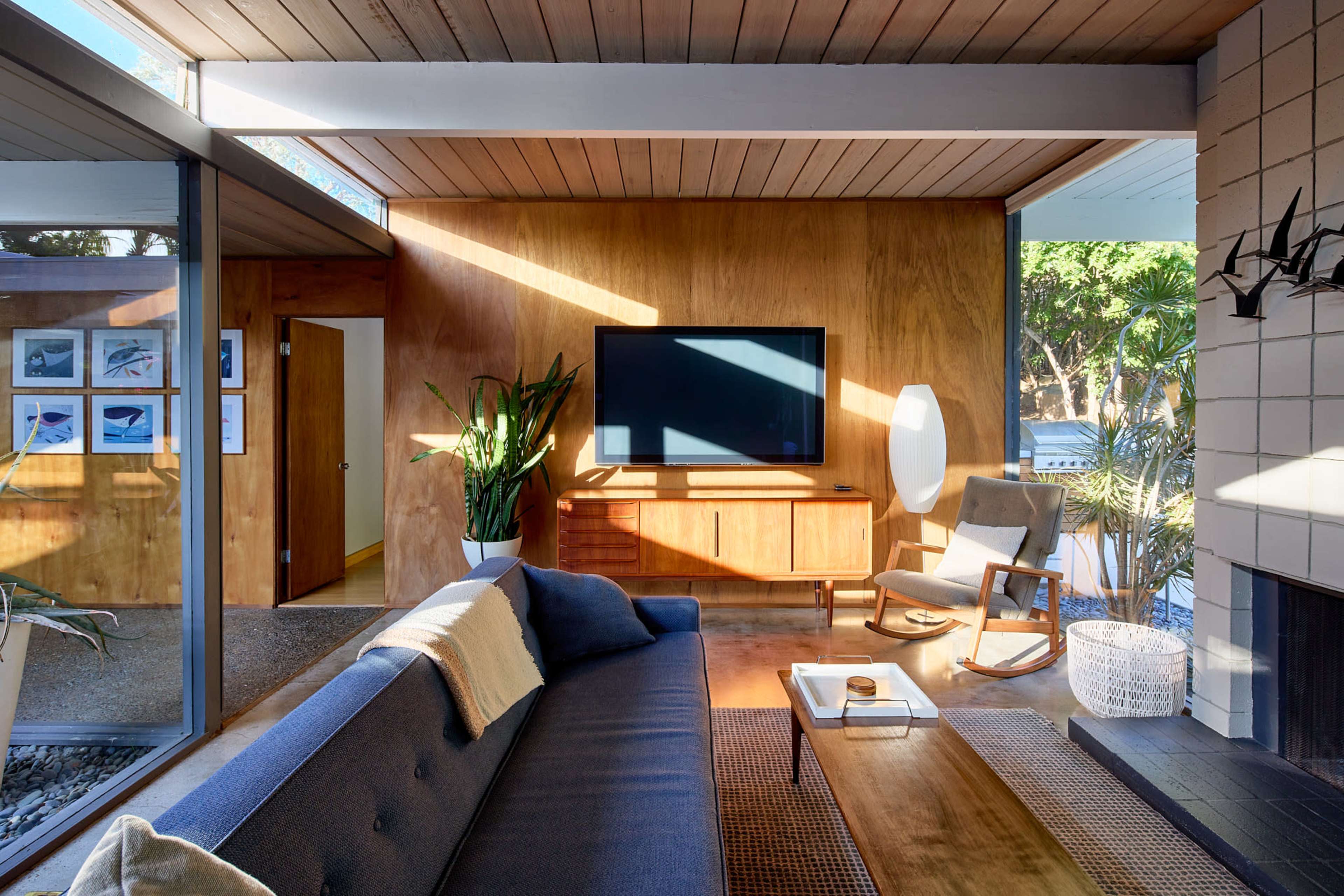 A mid-century modern living room features a blue sofa, a wooden media console with a TV, and large windows allowing natural light to illuminate the space.